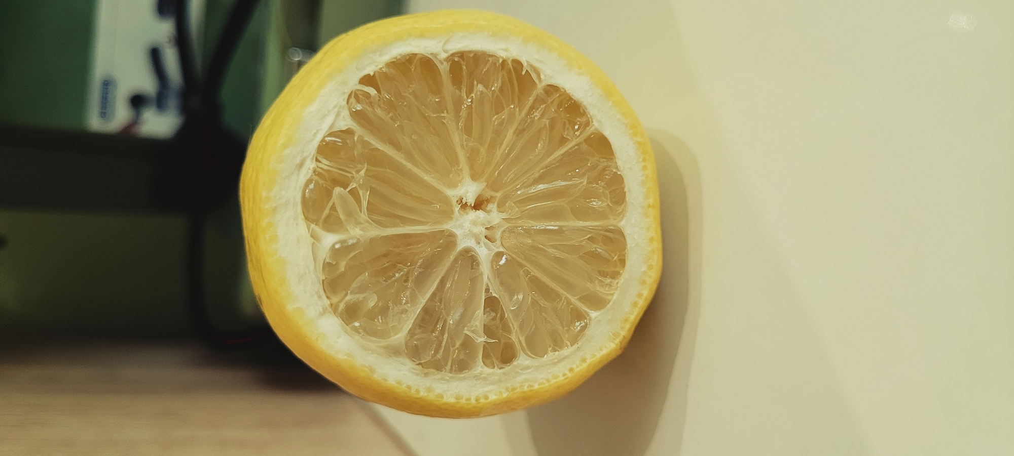 A close-up, eye-level, medium shot focuses on a halved lemon. The lemon is bright yellow on the exterior rind and the interior flesh is a translucent pale yellow, segmented into juicy sections. The background is blurred, with the left side featuring a vertical, light-brown, wooden surface, and the right side showing a dark green blurry background with a white electronic device. The lemon sits on a pale yellow surface. The lighting appears to be artificial, casting a soft glow. The image is a still-life, with no people or actions depicted.