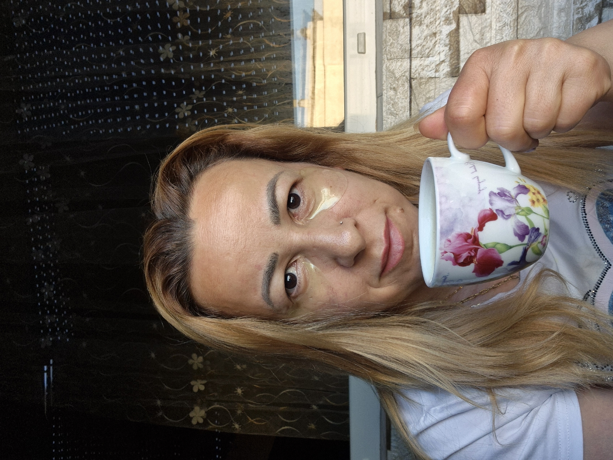 A woman is holding a floral patterned mug in her right hand. She has blonde hair and is wearing a white t-shirt. She is also wearing under-eye patches. The background appears to be a brick wall and a window with a patterned curtain. The image is taken outdoors, possibly on a balcony or patio. The lighting suggests it is daytime.