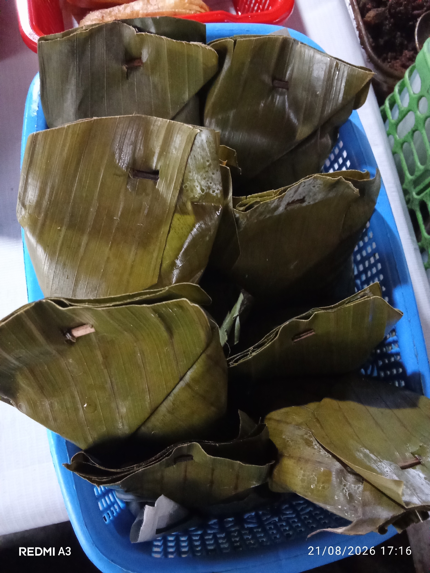 A close-up, overhead shot shows several food items wrapped in large green banana leaves, presented in a blue plastic basket. The food parcels are various sizes and shapes, some triangular and others more rectangular, secured with small pieces of wood or bamboo skewers. The banana leaves are textured with prominent veins and appear slightly moist, suggesting freshness or that the food is steamed. The blue basket has a woven pattern with visible holes. In the upper background, a red plastic container is partially visible, holding what appears to be more food. To the right, a green plastic container and some dark, granular food are also discernible. The foreground is dominated by the banana leaf-wrapped food. The image is marked with the text "REDMI A3" in the bottom left and "21/08/2026 17:16" in the bottom right, indicating the device used for capture and the date and time. The scene appears to be an indoor setting, possibly a market stall or a home kitchen in Purbayan, Indonesia, given the context of the food presentation.