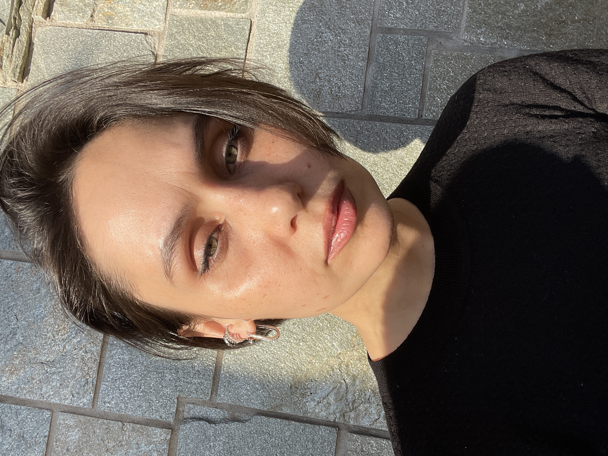 The image displays a close-up portrait of a young individual, rotated 90 degrees clockwise from a typical upright orientation, appearing to be lying down or leaning back with their head tilted to the left. The subject has short, dark brown hair, casually styled and slightly parted, with some strands framing the face. Their skin appears fair with some freckles visible on the nose and cheeks. The individual's eyes are open wide, looking directly at the camera, and appear to be a light color, possibly green or hazel. Their eyebrows are dark and well-defined. The lips are full, glossy, and a natural pink or nude shade, slightly parted.
The lighting is characterized by strong, direct sunlight hitting the left side of the subject's face (from the viewer's perspective), creating prominent highlights on the forehead, cheekbones, nose, and lips, which lend a dewy or oily sheen to the skin. The right side of the face is partially obscured by shadows cast by the nose and cheekbone, and a significant portion of the body is also in deep shadow.
The individual is wearing a black top, which appears to be made of a textured or knitted fabric with a soft, matte finish. A silver-colored earring, shaped like a miniature carabiner clip, is visible on the earlobe on the left side of the frame.
The background consists of a paved or tiled surface, likely an outdoor area, made of rectangular stone or concrete slabs in various shades of grey and beige. The sunlight casts sharp, distinct shadows onto this surface, including a large, rounded shadow that falls partly on the individual's clothing and the ground, suggesting an object (perhaps the photographer or a tree) is casting a shadow nearby. There is no visible text in the image.