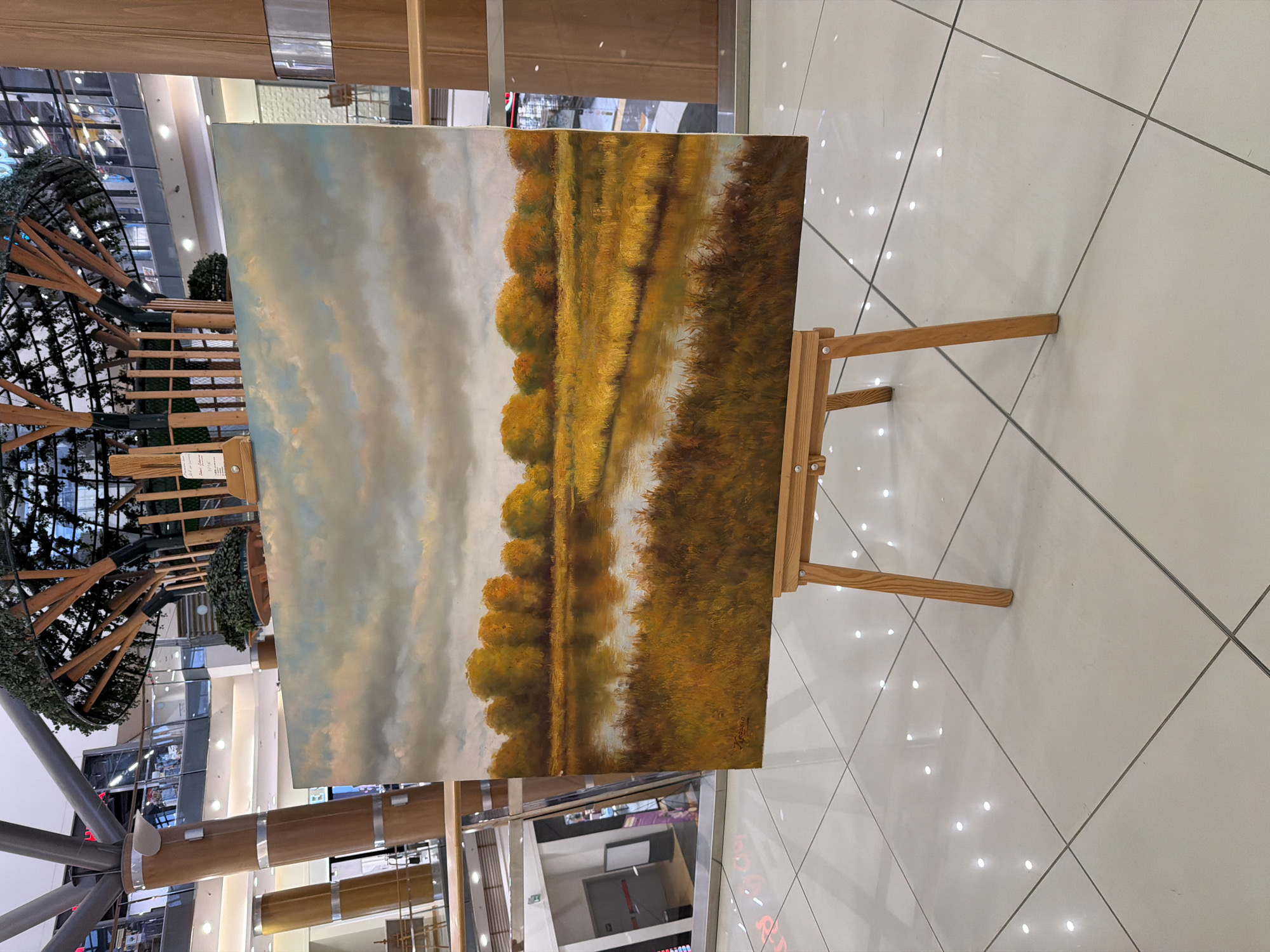The image displays a landscape painting presented on a wooden easel within a modern, multi-story indoor commercial building. The setting features a polished light-colored tiled floor reflecting bright overhead lights, large structural columns (some clad in wood with metallic bands, others appearing metallic), and glass railings delineating upper levels where shops are visible. A large, decorative tree-like structure constructed with wooden beams and artificial foliage occupies the central background.
The painting, positioned vertically on a light-toned wooden tripod easel, depicts an autumnal natural scene. It features a calm body of water, likely a river or stream, bordered by dense golden-brown grasses and reeds in the foreground and along the near bank. A line of trees with vibrant yellow, orange, and golden-green foliage stretches along the far bank. The sky above is overcast, with shades of grey and muted blue, showing subtle hints of pink or orange light. Reflections of the sky and trees are visible on the water's surface. A signature is discernible in the bottom left corner of the painting.
No individuals are present in the scene. A small information stand is partially visible behind the painting. A large reflective surface on the left side of the frame shows a partial reflection of an emergency exit sign and a doorway.
