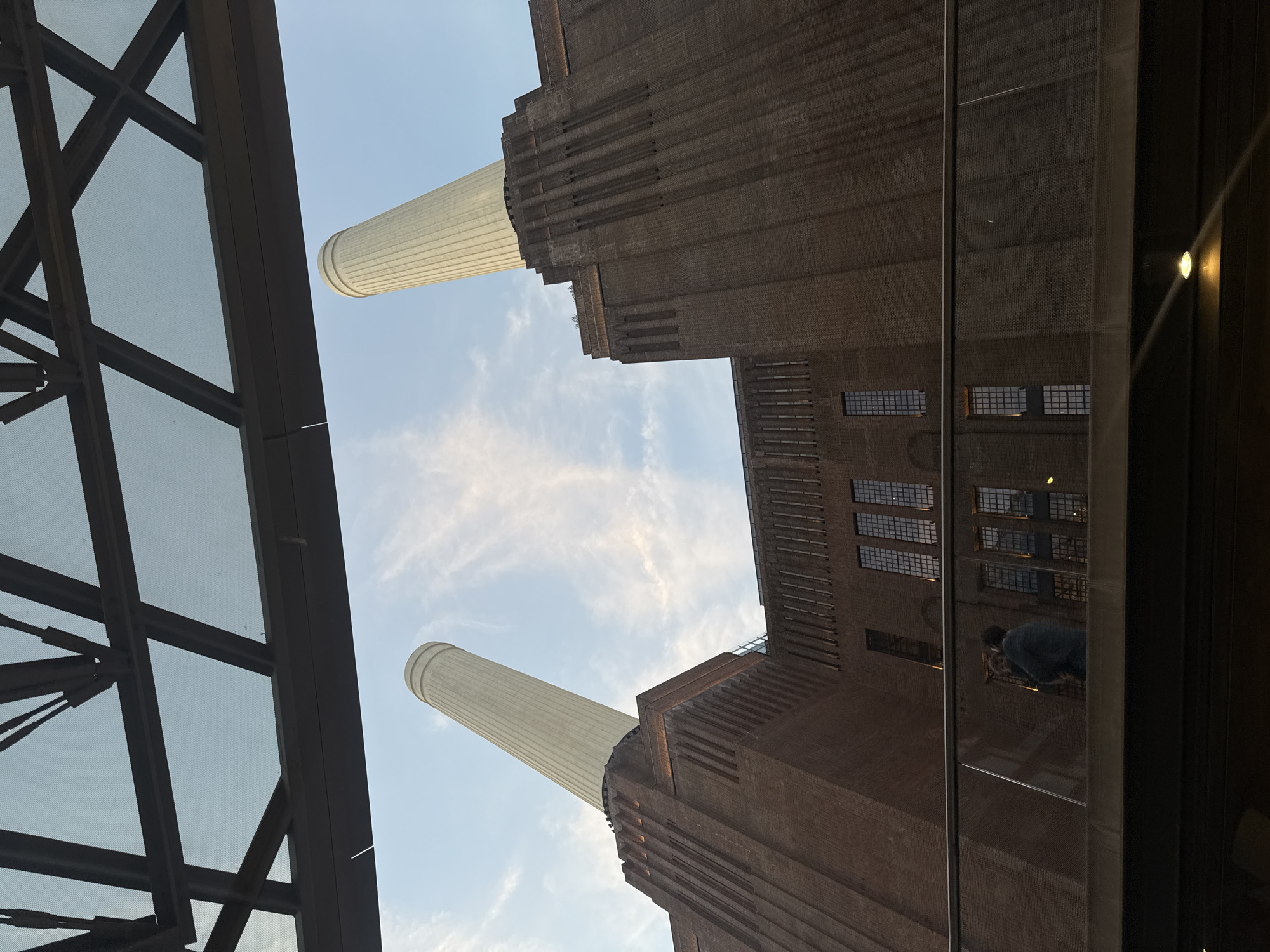 The image is taken from a low angle, looking up at the Battersea Power Station in London, United Kingdom. The sky is visible between two of the four iconic chimneys. A portion of a building's steel and glass structure appears on the left side of the frame, providing an unusual framing. The power station's brick facade is detailed with multiple windows, creating a symmetrical pattern. There is a reflection of a person or object visible in the glass structure in the bottom-right corner of the image.