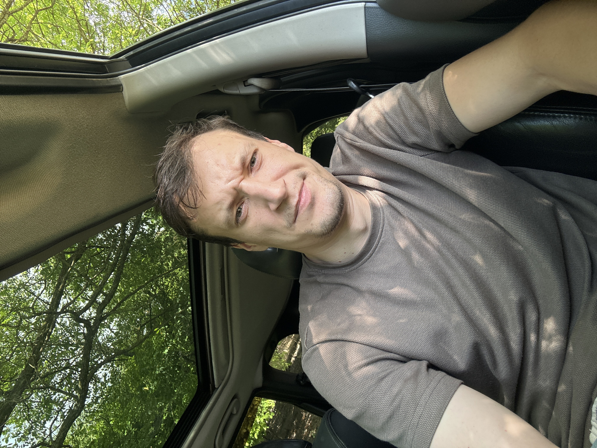 A man is sitting in the driver's seat of a car, looking at the camera with a furrowed brow. Sunlight streams through the open sunroof, dappling the man's face and grey t-shirt. The interior of the car, with black leather seats and grey headliner, is visible. Outside, green trees and foliage suggest a rural or wooded location. The image appears to be a selfie taken on a sunny day.
