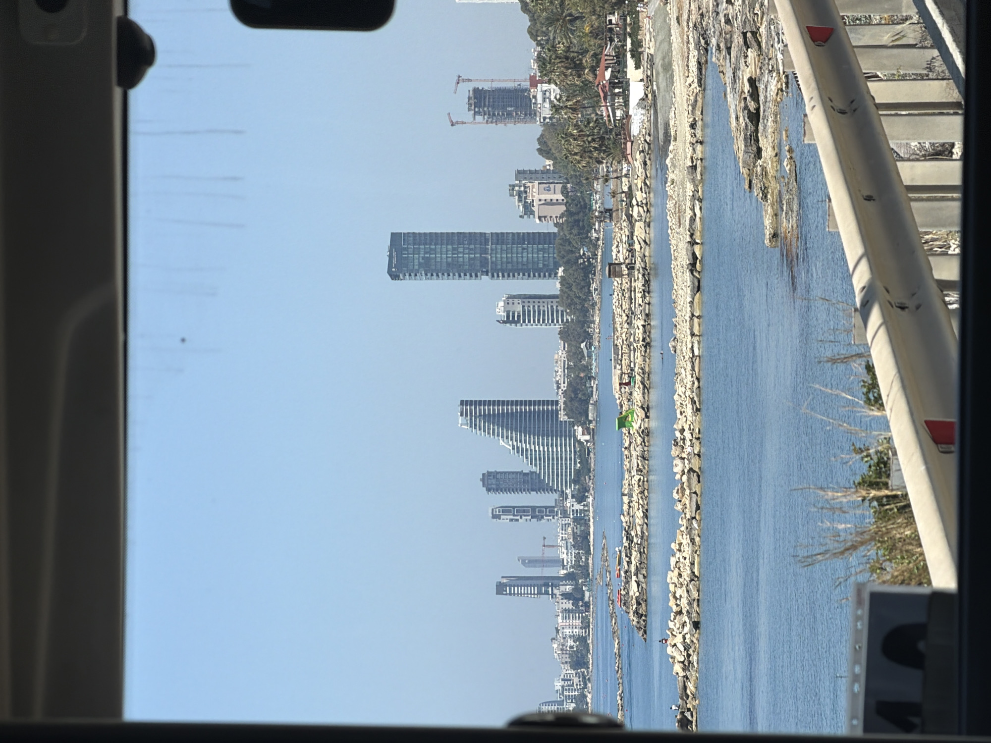 The image depicts a view from inside a vehicle, possibly a bus or car, looking towards the coastline of Agios Tychonas Community, Cyprus. The skyline is marked by several high-rise buildings, including a distinctive terraced building, under a clear blue sky. Between the vehicle's interior and the buildings is a body of water, separated from the urban area by a rocky shoreline. A portion of the vehicle's interior and window frame is visible at the edges of the image. A guardrail is visible through the window.