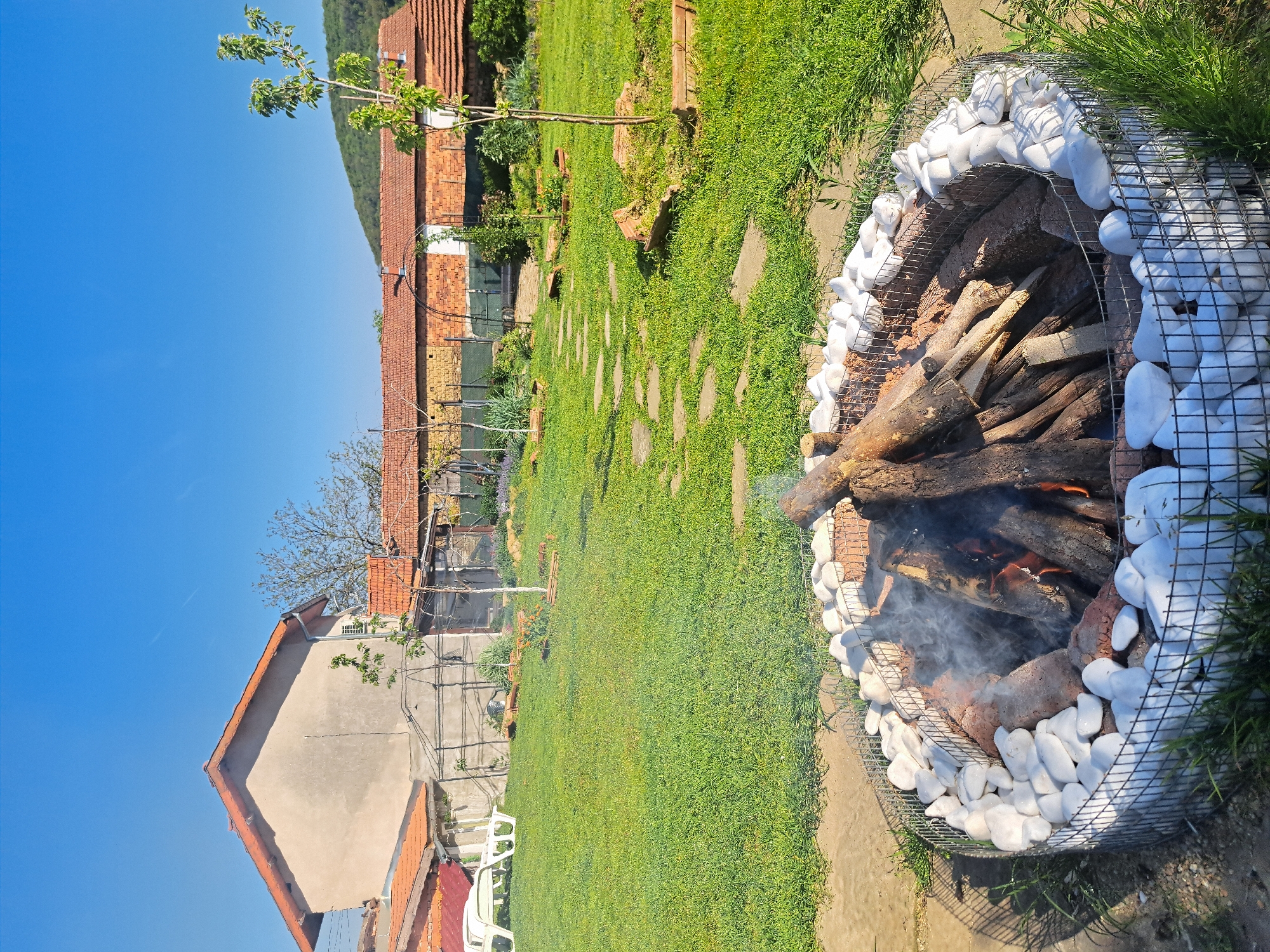 The image depicts a ground-level view of a yard in Lisets, Bulgaria. A circular fire pit constructed of wire mesh and lined with white stones contains burning logs and produces smoke. Behind the fire pit, a neatly maintained lawn leads toward buildings with red tile roofs. A stone pathway meanders through the grass. The sky is clear and blue. The buildings appear to be residential structures. White plastic chairs are visible near the base of one of the buildings.