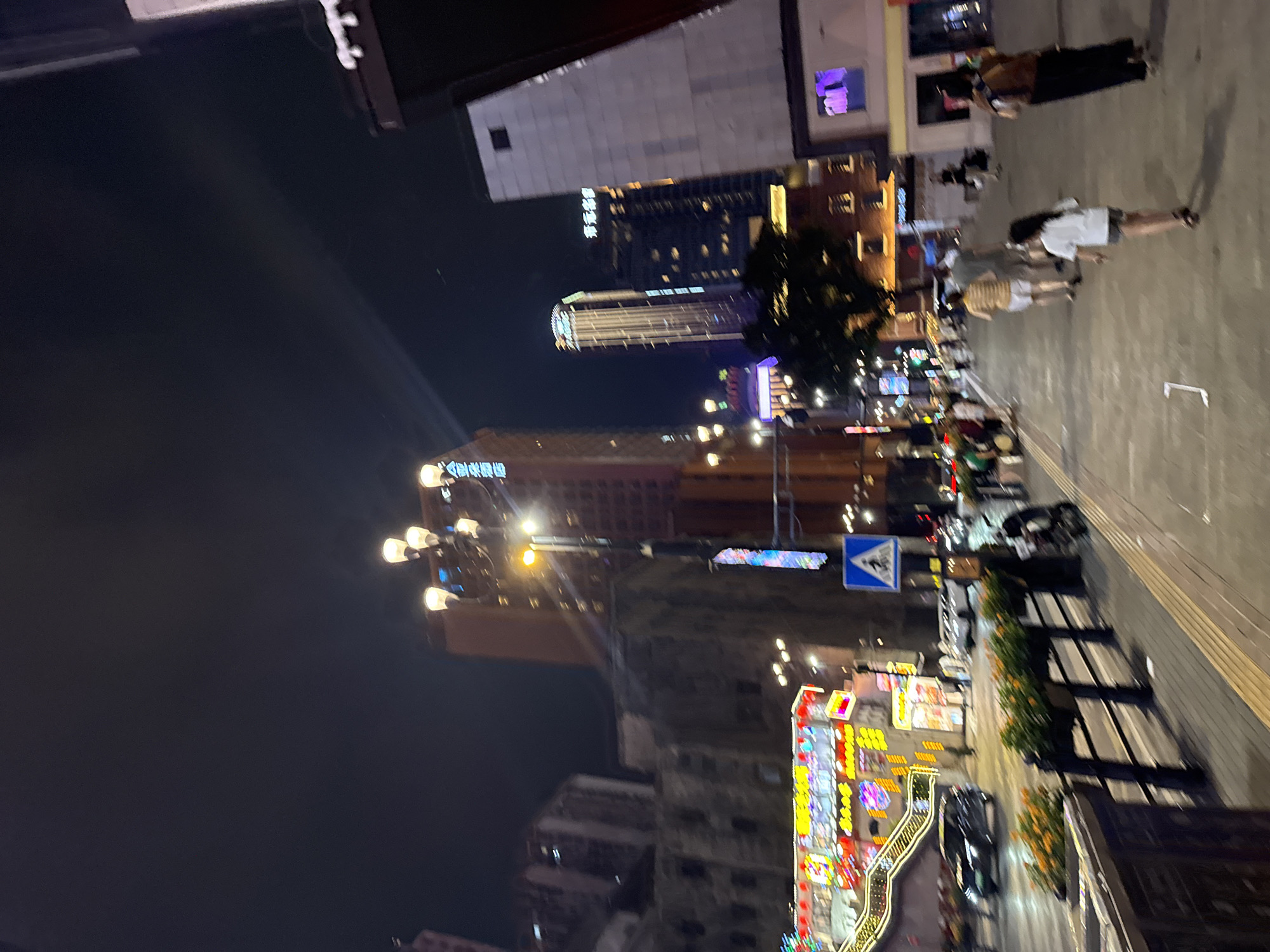 The image shows a nighttime street scene in Yuzhong District, China, likely a commercial area. A wide paved sidewalk is in the foreground, with a black metal railing and planters containing orange flowers and green foliage separating it from a street with car traffic. Several people are walking on the sidewalk. To the left, a brightly lit building with colorful signage is visible, featuring red lanterns and what appear to be illuminated stairs or ramps. Further in the background, tall buildings of varying architectural styles are illuminated against the dark sky. One prominent building is a slender skyscraper with vertical lighting. Streetlights are present, illuminating the scene with a warm glow. A pedestrian crossing sign is visible on the right side of the sidewalk. The overall impression is that of a busy urban environment at night.