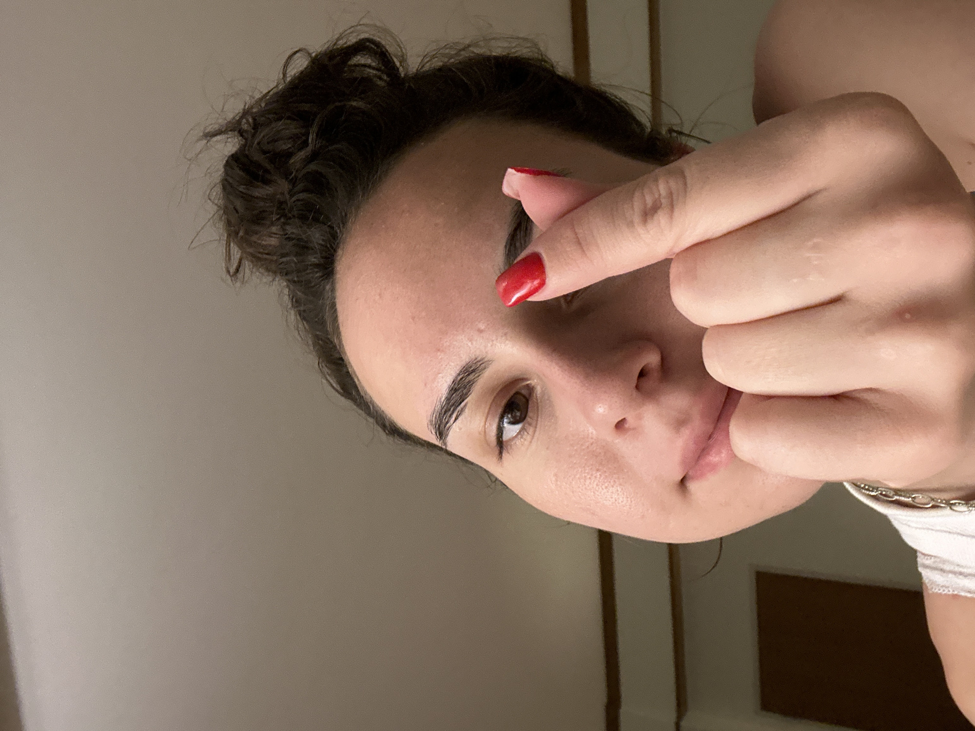 A woman with dark hair in a messy bun is close to the camera, holding up her hand with red nail polish, touching her forehead. Her face is slightly tilted, with her eye looking directly at the camera. The background is blurry and appears to be a wall, possibly a room. A silver chain necklace is visible on her neck. The image was not taken in Pattaya City, Thailand.