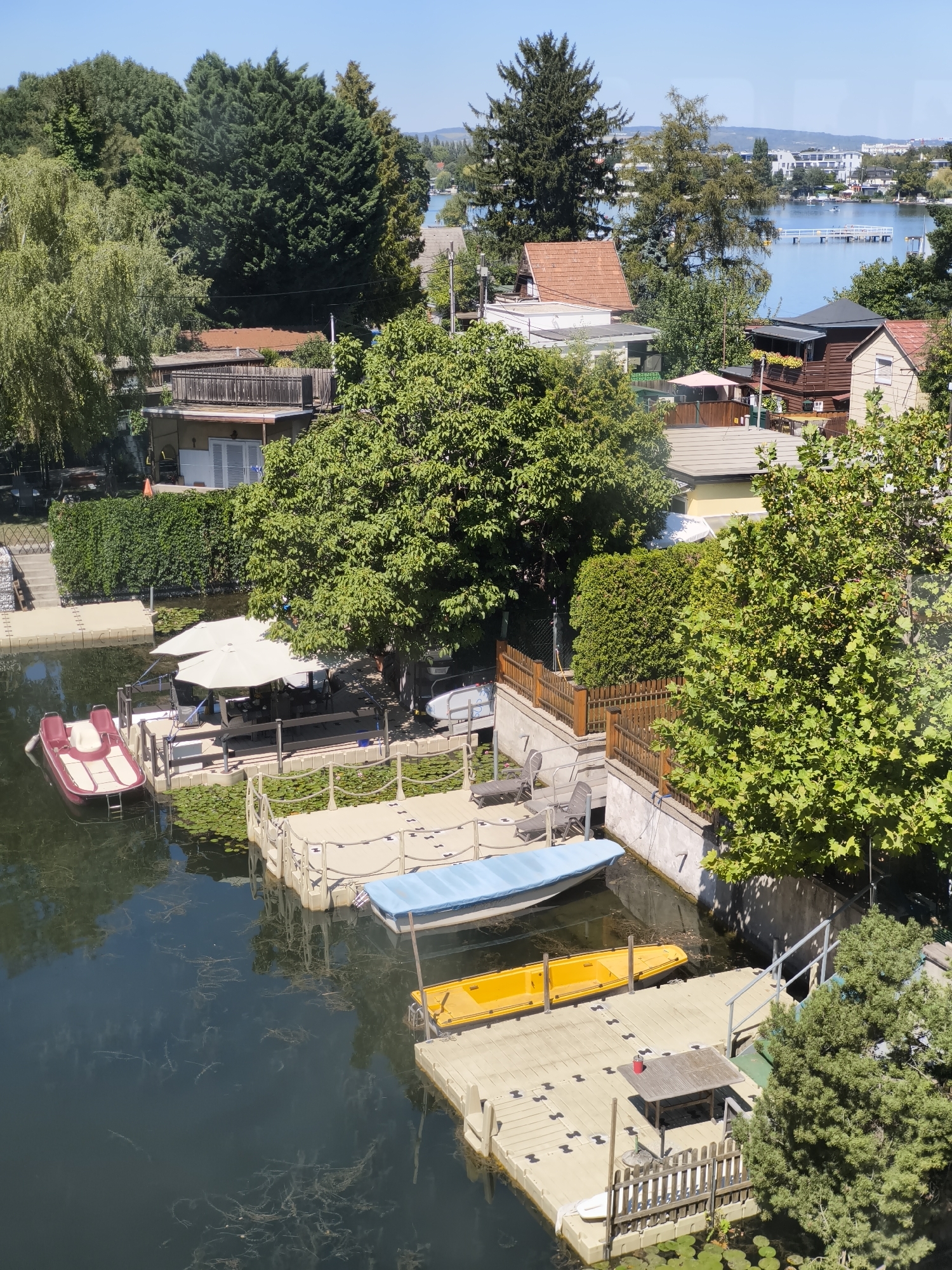 This is an aerial, eye-level, medium shot taken on a sunny day. The scene depicts a waterfront area with several houses and boats. In the foreground, a body of water with lily pads is visible, with a brown and white pedal boat docked on a floating wooden platform. Adjacent to this, a blue-covered boat and a yellow kayak are also docked. Further back, a house with a red tile roof and a wooden balcony is seen, with trees and vegetation surrounding it. In the background, a wider body of water, likely a lake or river, stretches out, with buildings and trees lining the opposite shore. The overall impression is one of a peaceful residential area by the water. While the image is visually detailed, the precise location within Vienna, Austria, cannot be determined solely from the visual information present.
