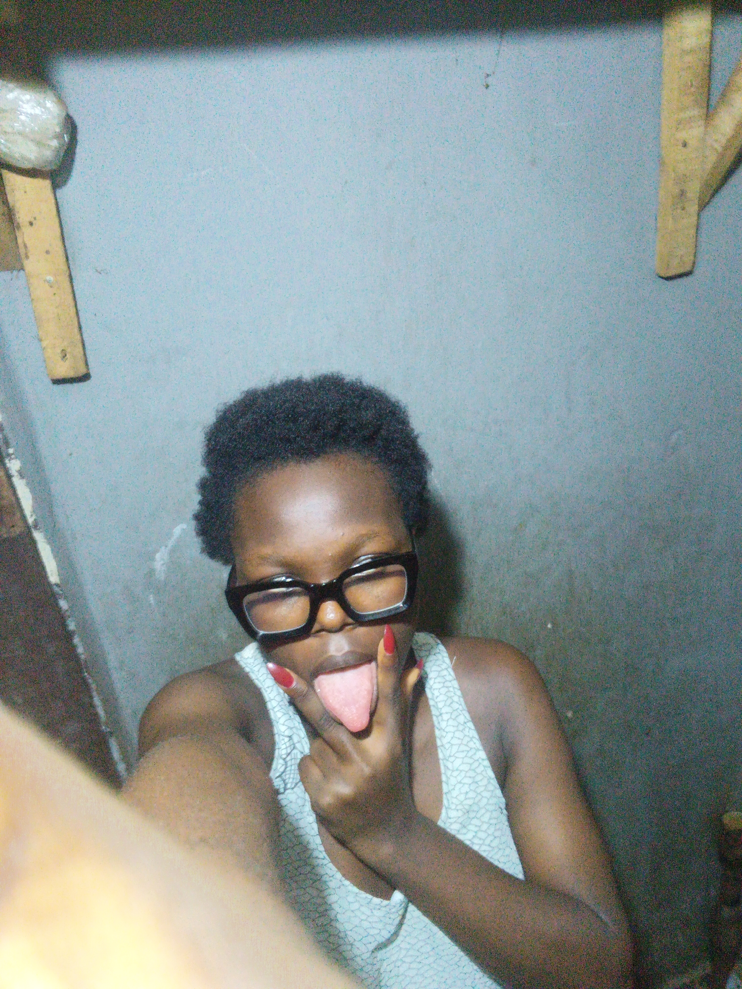 The media file is a medium shot selfie of a young Black woman in a dimly lit, possibly indoor setting. She is wearing glasses with thick black frames and a light-colored patterned tank top. Her mouth is open, and her tongue is sticking out, with one hand raised, index finger pointing towards her mouth, and her tongue sticking out. Her fingernails are painted red. The background appears to be a textured wall, with some wooden elements visible in the upper corners. There is also a hint of what looks like a shelf or ledge on the left side of the frame, with a wrapped object on it. The lighting is uneven, creating shadows and highlights on the subject and the background. The overall mood appears playful or provocative due to the facial expression and hand gesture. Based on the provided location context of Onitsha, Nigeria, this could be a personal snapshot taken in a residential or casual environment. No visible text is present in the image.
