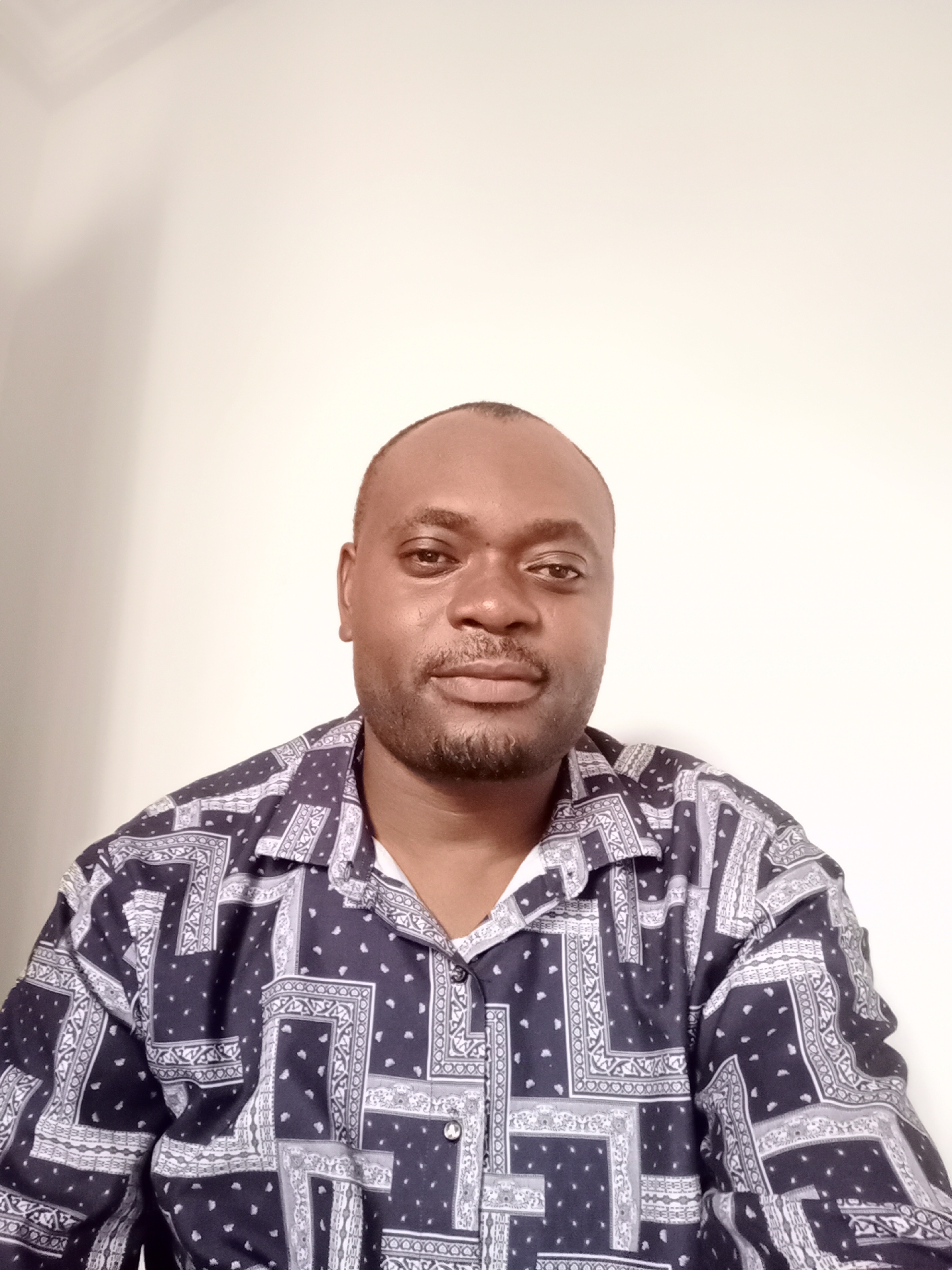 A medium shot depicts a man against a plain, light background. The man is dark-skinned with a shaved head and a short, well-groomed beard. He is wearing a button-down shirt with a complex black and white paisley-like pattern. His gaze is directed towards the camera. There are no other discernible people, subjects, or objects in the frame. The environment is devoid of distinguishing features. The image does not provide any location information related to the city of Gwarinpa, Nigeria, nor does it offer any clues to make such an association.