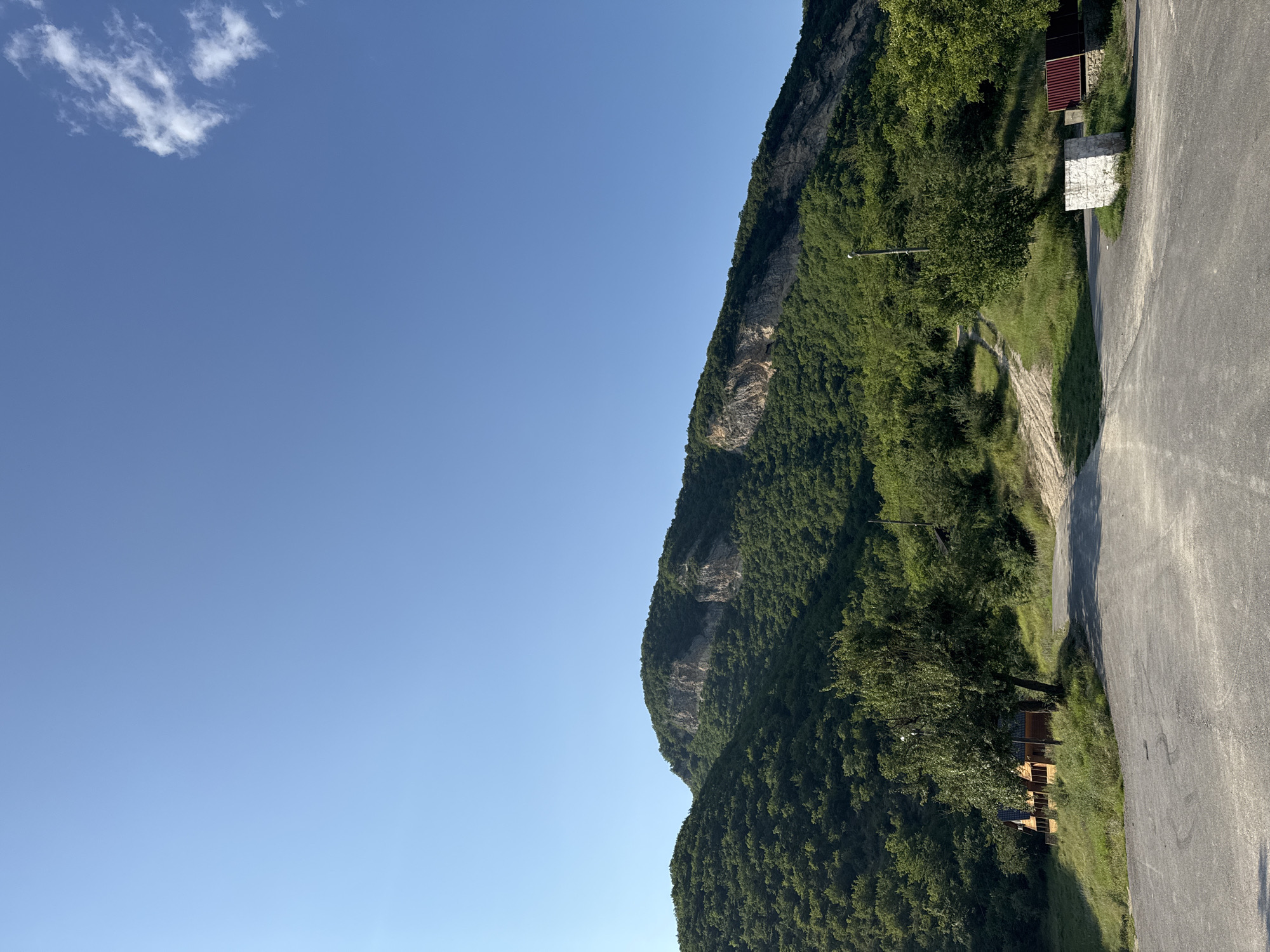 The image shows a scenic view of a mountain range covered in lush green trees, with a clear blue sky above. In the foreground, there is an asphalt road leading towards the mountains. To the left of the road, a small wooden house with a dark roof is nestled on a grassy slope, with a large tree beside it. Further to the right, there are some metal fences and a white stone structure. A dirt path is visible winding up the side of the mountain, disappearing into the trees. The overall scene is peaceful and natural, suggesting a rural or mountainous location.