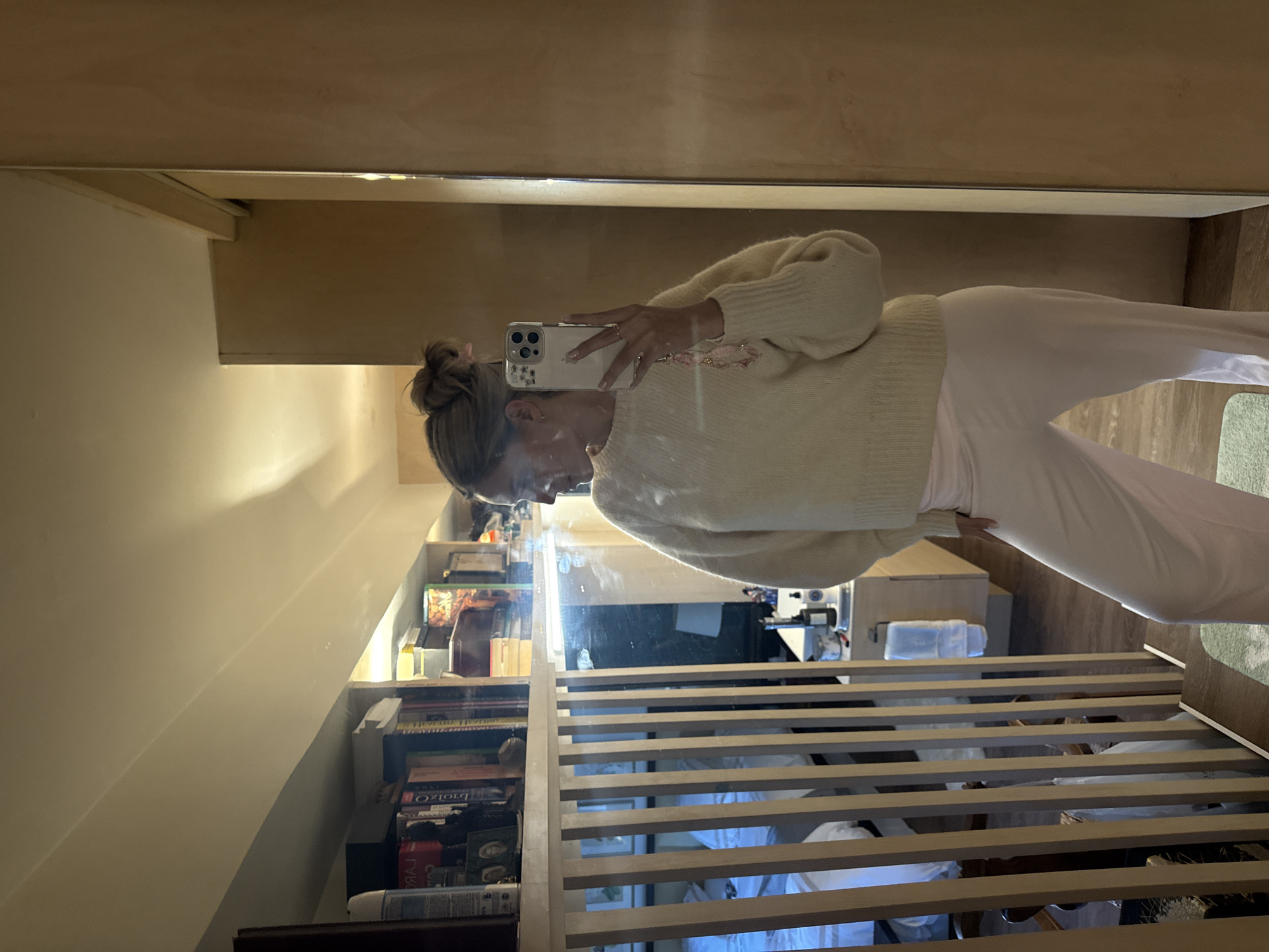 A woman with blonde hair pulled into a bun is taking a mirror selfie. She is wearing a light yellow, fuzzy sweater and pale pink, satin pants. Her left hand is holding a smartphone, and her right hand is resting on her hip. She has light makeup on, and a ring is visible on her left hand. In the background, a shelf filled with books and decorative items is visible. A bed is also reflected in the mirror, along with a wooden partition. The room appears to be a bedroom or dressing area, with warm lighting. The image does not contain information to determine a location in Ciudad de México, Mexico.