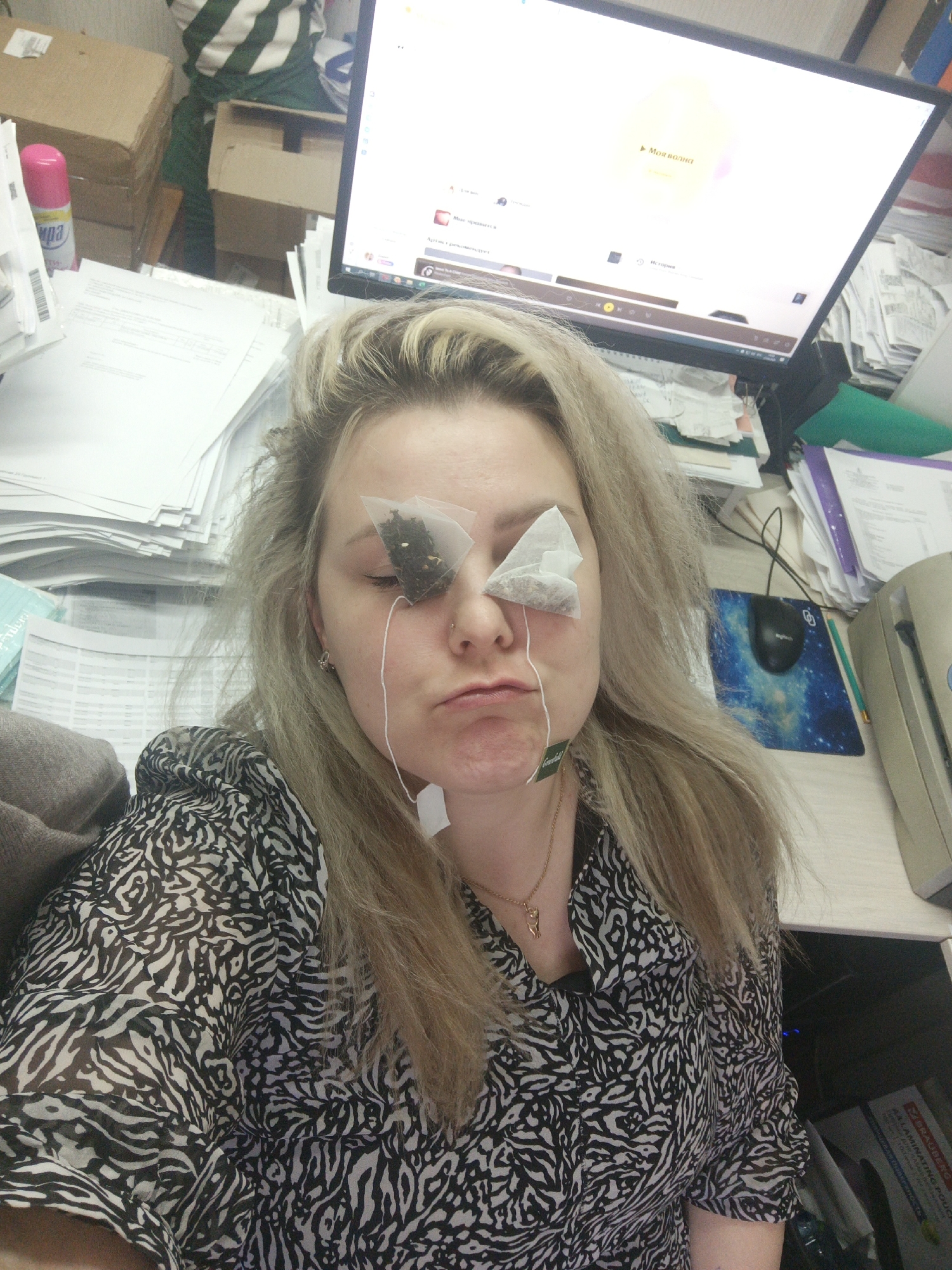 A woman with blonde hair is seated at a cluttered desk in Noviy, Russia. She has two tea bags placed over her eyes, with the strings hanging down. She wears a black and white patterned blouse and a gold necklace. The desk is covered in stacks of paper, boxes, office supplies, a computer monitor, a mouse pad with a galaxy print, and a laminating pouch. The computer monitor displays a webpage in Russian. The environment appears to be an office setting.
