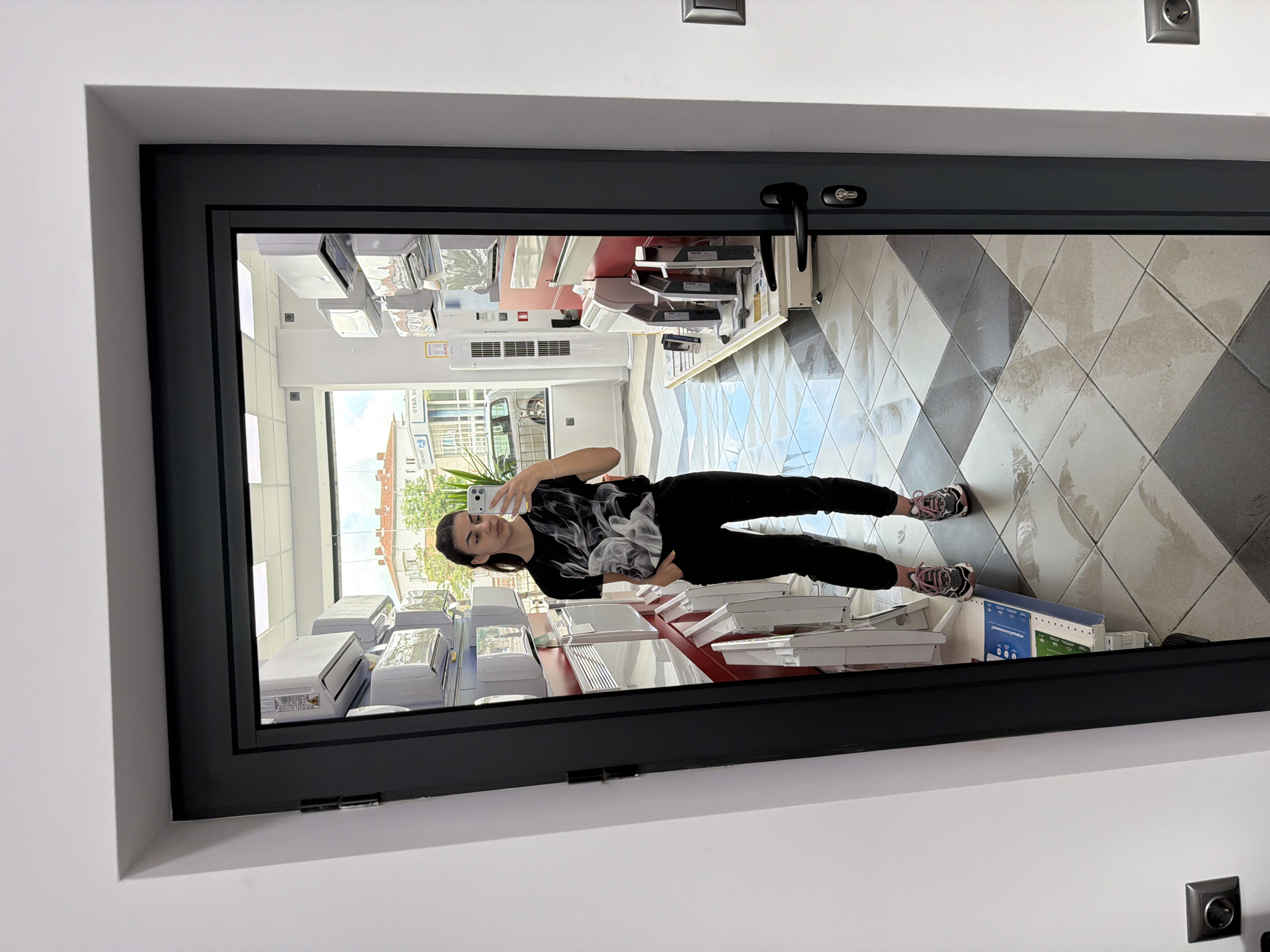 A woman stands in a commercial establishment. The woman is taking a photograph of herself with a smartphone. The building appears to be a retail store that sells or displays air conditioning units. The floor is tiled in a diamond pattern with alternating light and dark colors. Based on visible signage in the reflection, the store appears to be located in Gabrovo, Bulgaria.