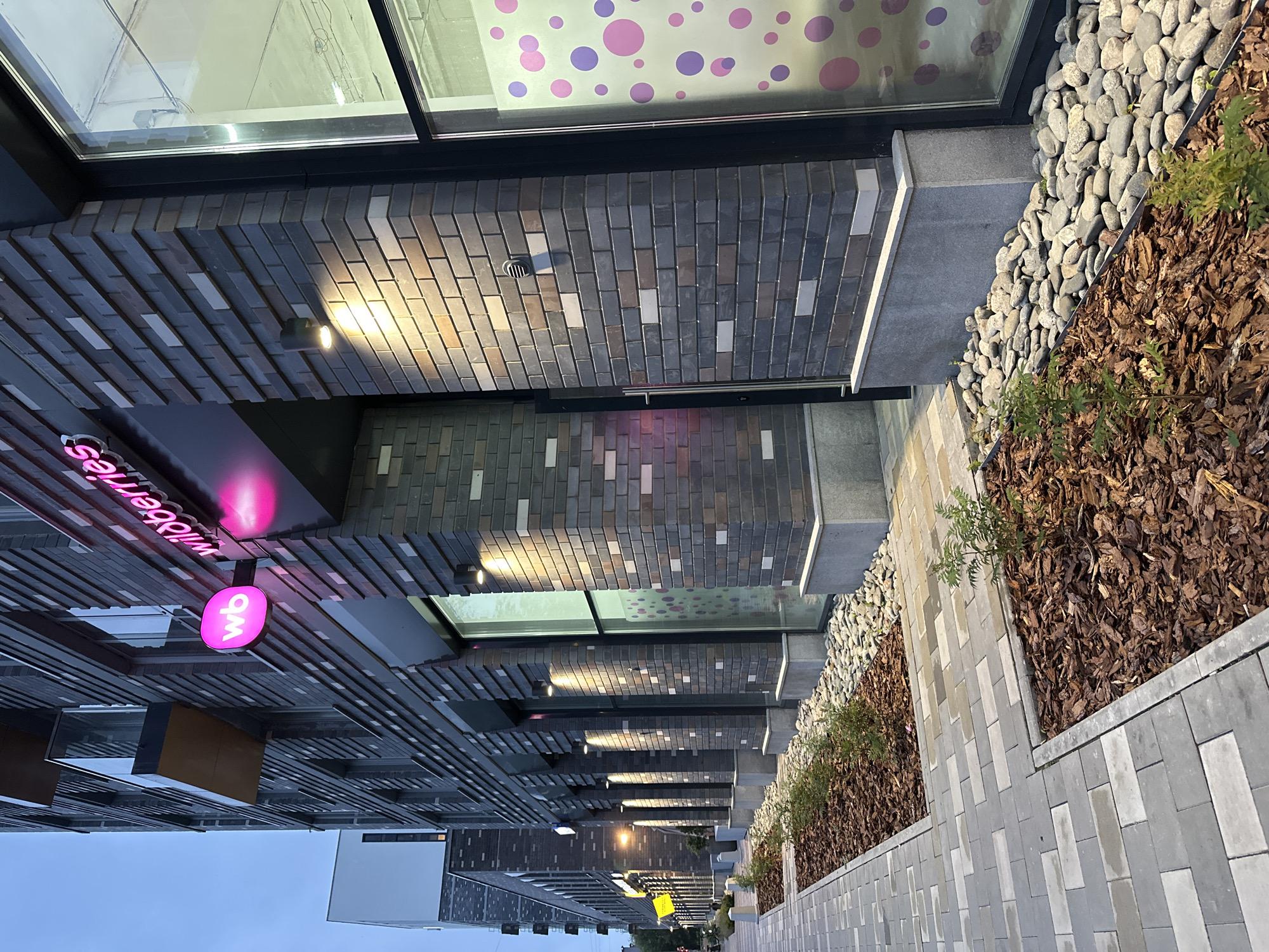 The image shows the exterior of a modern commercial building in Yekaterinburg, Russia. The building features a facade constructed with dark grey and lighter grey bricks, with some rows featuring white bricks. A prominent sign with "wb" in a glowing pink circle and the word "wildberries" in pink neon letters is mounted on the building's facade. Several windows are visible, one of which has a decorative pattern of pink and purple circles. Exterior lighting fixtures cast warm light onto the brickwork. A paved sidewalk with a patterned design runs alongside the building, bordering planter beds filled with mulch and small green plants, and also with river stones. In the background, other modern buildings and a cloudy sky are visible. No people are present in the image.