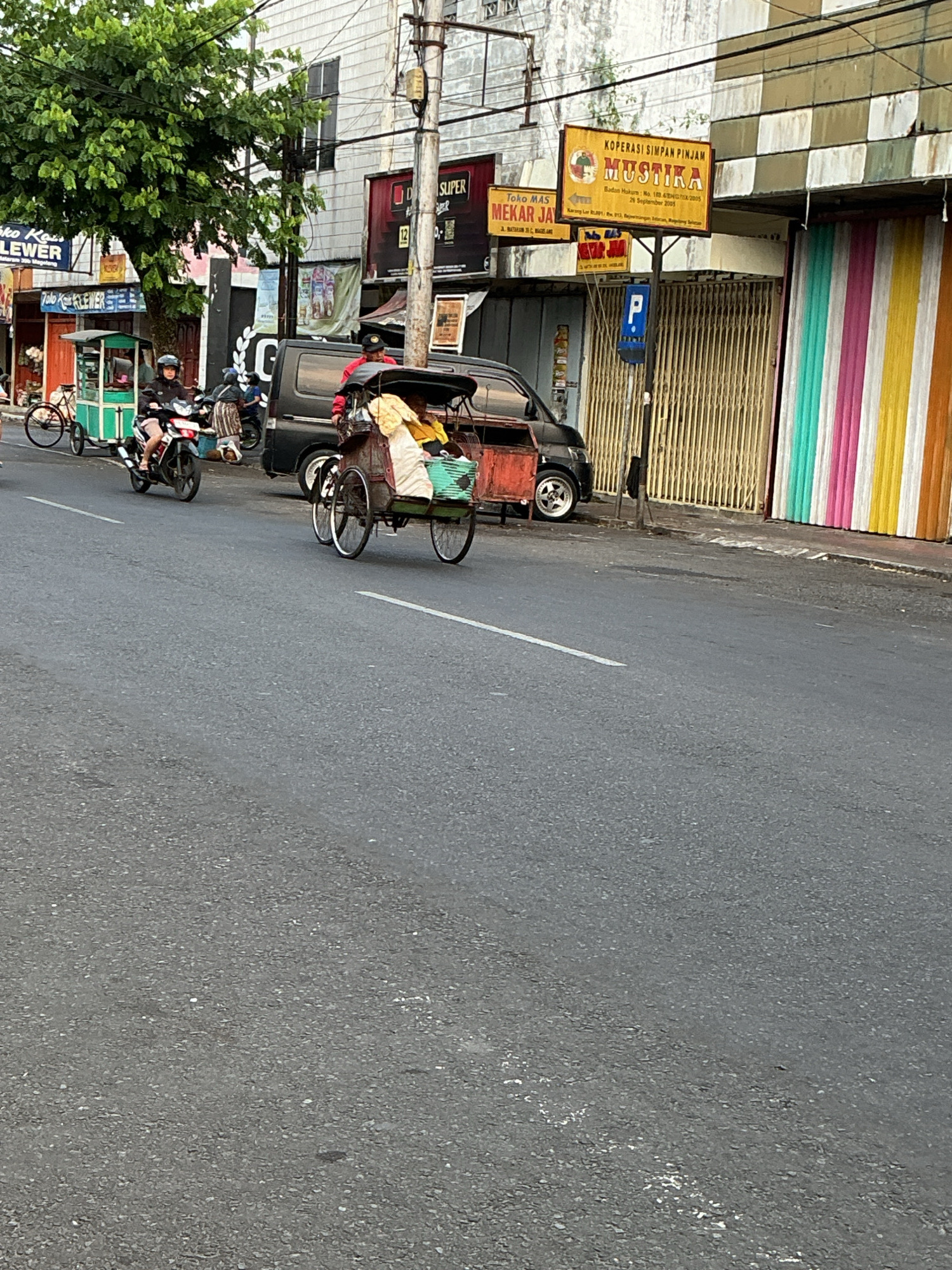 This image captures a street scene in Magelang, Indonesia, focusing on vehicular traffic and storefronts. In the foreground, a gray asphalt road shows white lane markings. A three-wheeled pedicab, with a red cargo box at the rear and two occupants, travels on the right side of the road. The pedicab is laden with what appears to be textiles or cloth. To the left of the pedicab, a dark minivan is parked. Further to the left, a person on a red motorcycle is seen riding past a green and white pushcart. Several buildings line the street. One building has a sign that reads "KOPSIMPINJAM MUSTIKA" with a logo of a traditional puppet figure. Adjacent to it is a storefront with a sign reading "Mekejay," featuring images of women. Another building displays a sign for "SUPER." The storefronts have a mix of closed metal shutters and colorful vertical striped panels. A mature tree with dense green foliage is visible on the left side of the image, casting shadows. Utility poles and wires are prominent above the buildings, indicating urban infrastructure. The overall impression is a snapshot of daily life and commerce on a street in Magelang.