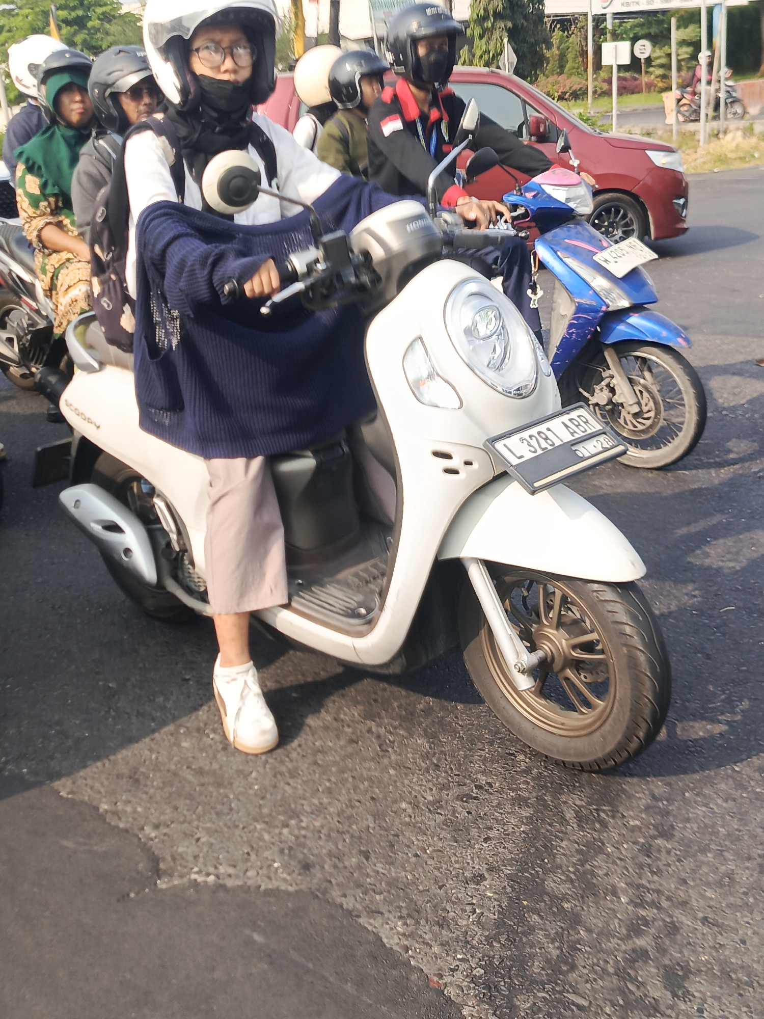 In Calukan RT 1 RW 5 Keboansikep, Indonesia, three people are riding on two motorcycles on a paved road. The foreground shows a white scooter with a female driver wearing a blue sweater, light-colored pants, and white shoes. She has glasses and is wearing a helmet and mask. A second person, also wearing a helmet and mask, is seated behind her on the same scooter. The scooter has a license plate that reads "L 3281 ABR". In the background, another motorcycle is visible with two people, one driving and one passenger, both wearing helmets. The driver of this second motorcycle is wearing a black jacket with red and white accents. A red car is also present in the background, and the scene appears to be during daylight with a clear sky. The road surface shows some patches of wear and tear.