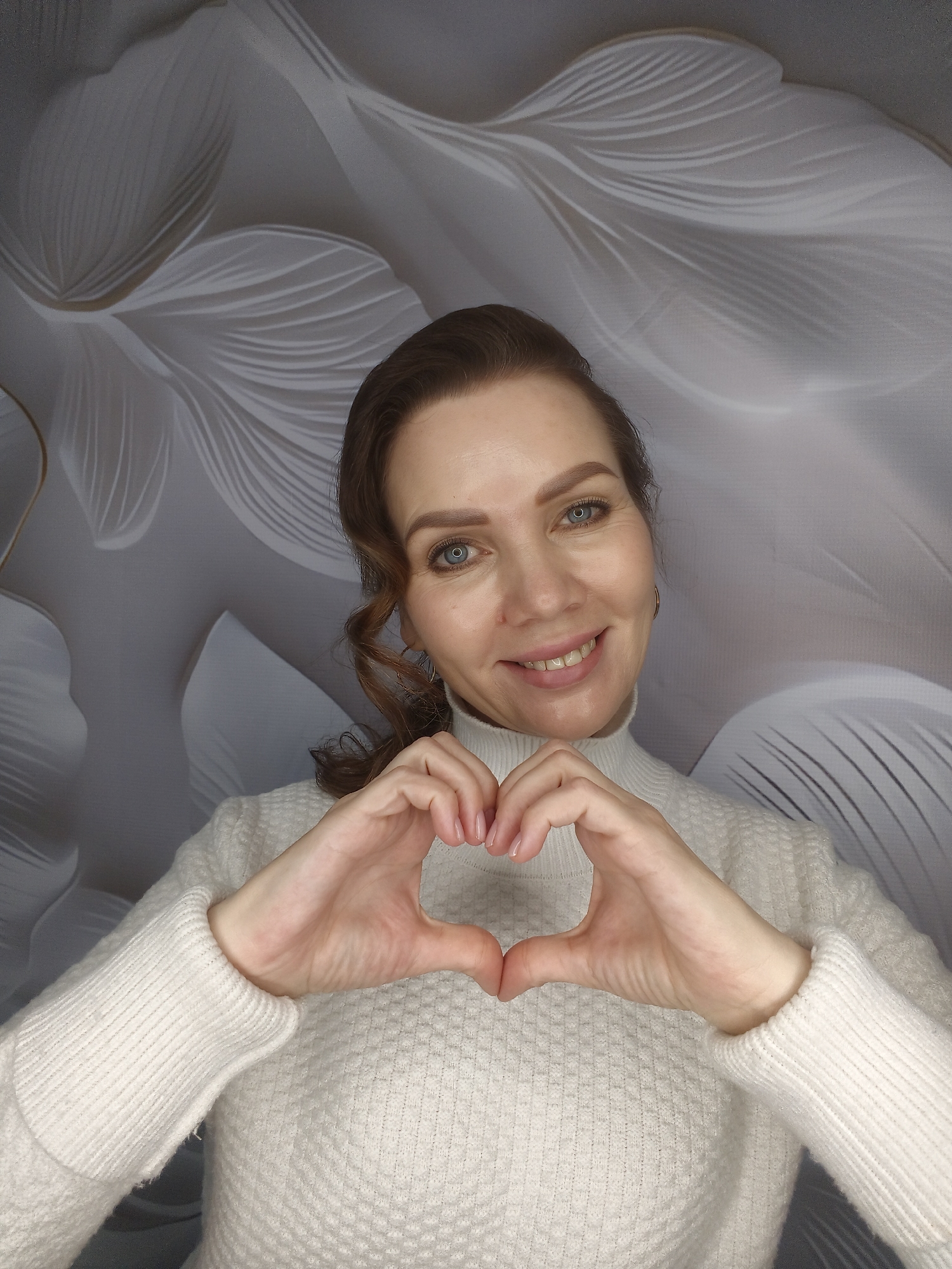 A woman with brown hair pulled back into a low ponytail is smiling at the camera while making a heart shape with her hands. She has fair skin, blue eyes, and is wearing a white turtleneck sweater. The background is a textured wallpaper with a large, abstract floral or feather pattern in shades of gray and white. The photograph appears to be taken indoors with soft, even lighting. The location is not identifiable from the image.