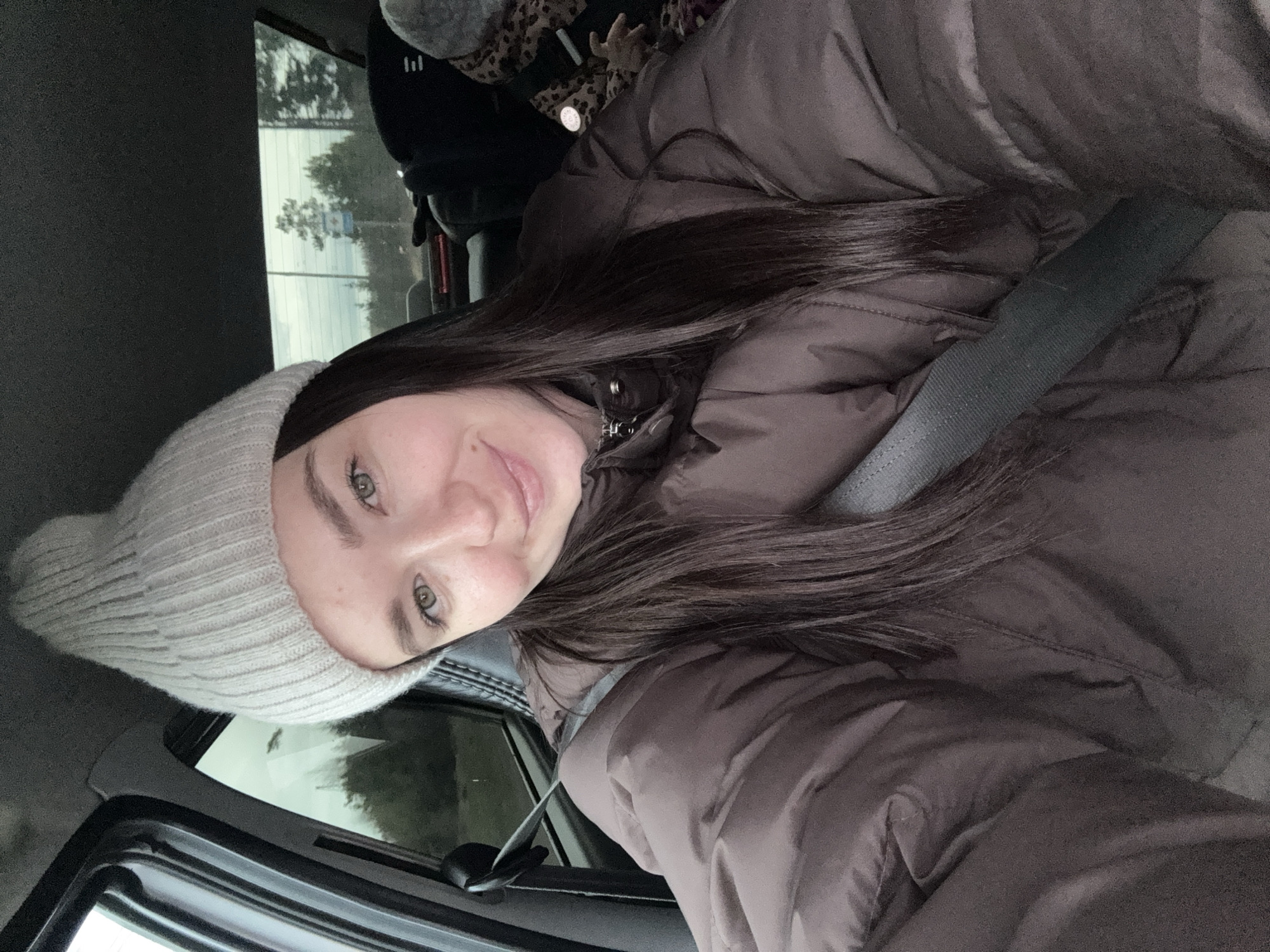 A young woman is captured in a close-up, likely a selfie, within a car interior. She has long, dark hair, light-colored eyes (appearing green or hazel), and a fair complexion, with a faint smile on her face. She is wearing a light-colored, possibly off-white or light grey, knitted beanie and a dark brown or charcoal puffer jacket. A seatbelt is visible across her chest, indicating she is a passenger.
Through the car's rear window, an overcast sky and scattered trees are visible, suggesting daytime with gloomy or cloudy weather. The interior of the car appears standard, with dark upholstery and window frames. In the background, behind the woman's head, hints of a leopard print fabric or accessory can be seen, along with a dark object that might be a bag or a headrest. The overall impression is that she is traveling in the car, potentially in Гаревая, Russia, based on the provided location context. No discernible text is visible in the image.