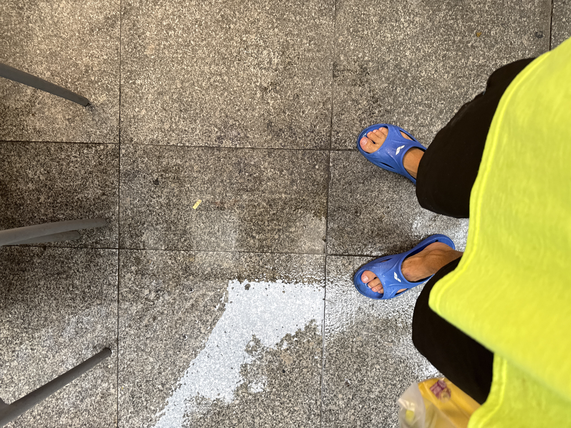 The image, taken from a high angle, shows two feet wearing blue slide sandals on a wet, grey speckled tile floor. The tiles appear to be granite. A puddle of water is visible on the left side of the frame. The person is wearing black pants and a bright yellow shirt or vest, the fabric of which fills the bottom of the frame. The legs of at least two grey metal chairs are visible at the top of the image. The scene is likely outdoors, possibly on a patio or sidewalk in Haizhu District, China, given the context.