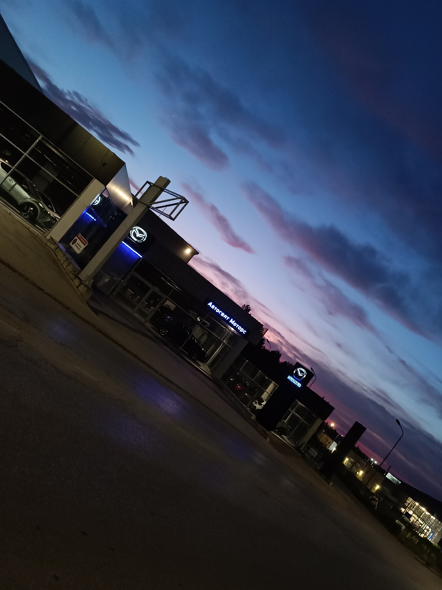 The media file presents an exterior view of a car dealership at dusk or early night, captured with a dynamic, tilted perspective. The primary subject is a modern building housing a Mazda dealership, prominently displaying illuminated "MAZDA" logos and the name "Автосвят Моторс" (Avtosvyat Motors) in Cyrillic script. The building features large glass windows, through which the interiors and some cars, likely new models, are visible. The architecture is contemporary, with sleek lines and bright blue neon lighting highlighting elements of the facade.
The setting appears to be along a street or commercial area, with an asphalt surface in the immediate foreground reflecting some of the neon light. No people are visible in the scene.
A notable detail is the sky, which transitions from a deep, dark blue at the top to vibrant purplish-pink and lighter blue hues closer to the horizon, indicating either the last vestiges of sunset or the early stages of twilight. Scattered clouds stretch across the sky, catching these twilight colors. The dealership's lights, including the illuminated signs, are brightly lit, contrasting with the darkening sky.
A red circular sign with white text, partially obscured, is visible on the left side of the building; it appears to be a warning or instruction sign, possibly reading "ВНИМАНИЕ" (Attention) and "ОГЛЕД" (Inspection). A street light can be seen further down the road on the right. The overall impression is one of quiet commerce as day transitions to night.