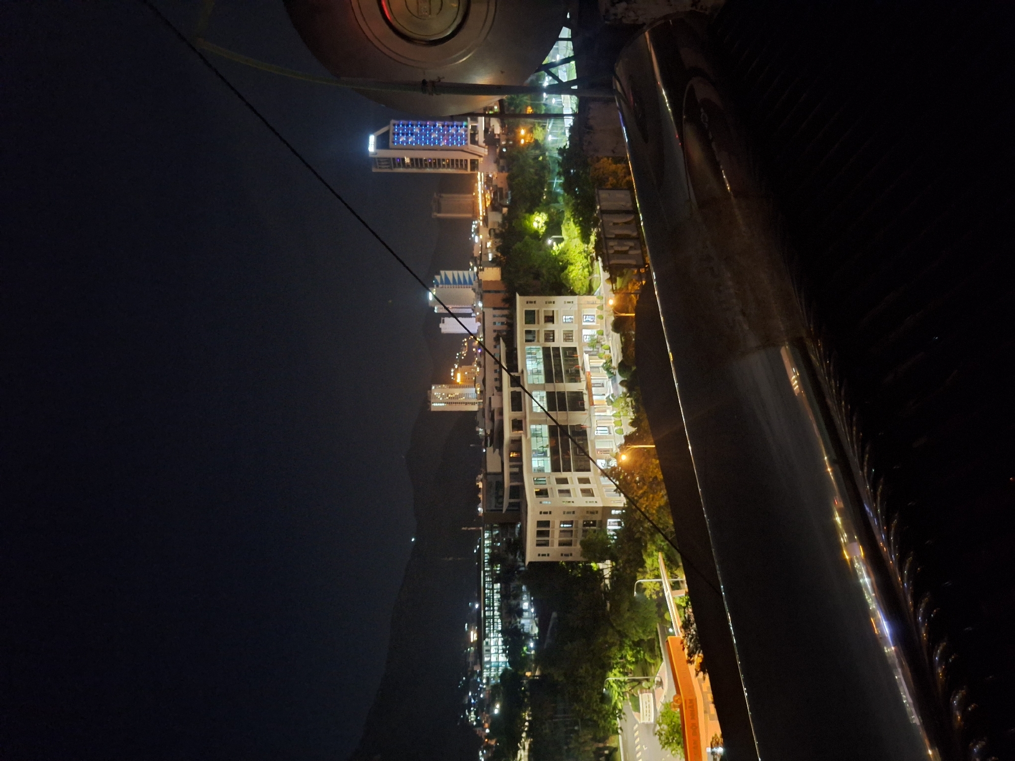 This is a nighttime photograph of a city skyline in Nha Trang, Vietnam. The image is taken from an elevated perspective, possibly from a building or a cable car, looking down and across at the city.
The foreground features a dark, indistinct object that appears to be part of a structure, possibly a railing or a part of the vehicle from which the photo is taken. A thin cable stretches diagonally across the frame from the upper left to the lower right.
The midground and background are dominated by the brightly lit city. Several multi-story buildings are visible, with lights on in many of the windows, suggesting it is a busy urban area. One tall building in the distance is illuminated with a pattern of blue lights. Green trees and foliage are interspersed among the buildings, indicating that the city has green spaces. Streetlights and lights from vehicles are also visible, creating a vibrant urban glow against the dark sky. In the far distance, dark hills or mountains are silhouetted against the night sky.
The time of day is clearly night, as indicated by the darkness of the sky and the prominence of artificial lighting. The weather appears to be clear, with no visible rain or fog. The overall impression is one of an active and well-lit city at night, with a mix of modern architecture and natural elements. There is no visible text in the image.