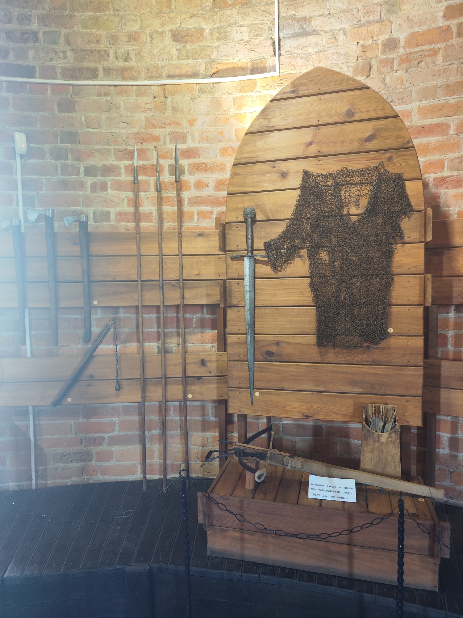 An exhibit of medieval weapons is displayed against a brick wall in Kamyanyets, Belarus. The display includes a chainmail shirt, a sword, several spears, two axes, and a flail. A crossbow is also visible in the foreground, resting on a wooden chest. A sign in both Russian and English reads "Don't touch the exhibits." The lighting is dim, with a spotlight highlighting the weapons.