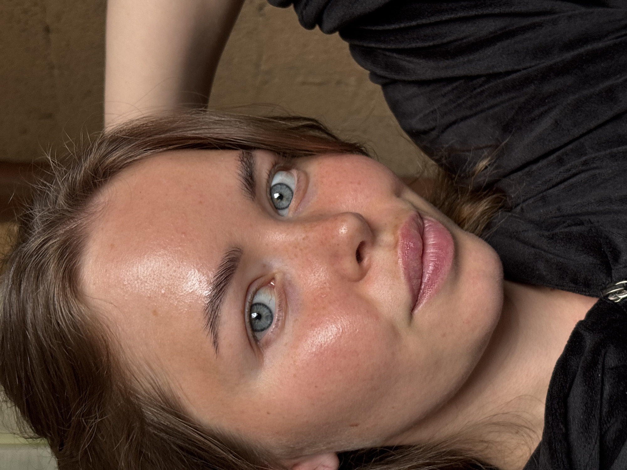A close-up image captures a young woman with light skin, freckles, and grey eyes, positioned at an angle. Her face is the primary subject, and she appears to be looking directly at the viewer. She has brown hair and is wearing a black velour top. The background is a neutral, slightly textured surface. No specific location in Русский Вожой, Russia, is identifiable from the image.