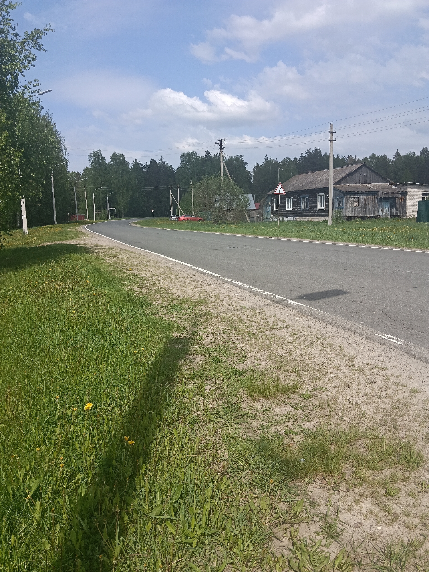 The image depicts a road in the city of Красный Маяк, Russia. The road curves gently to the right and has a white line marking the edge. The side of the road is grassy with some dandelions. On the right side of the road is a weathered wooden house with a small annex. There is a road sign next to the house. Utility poles run along the side of the road. A forest is visible in the background. The sky is blue with scattered clouds. A red car is parked further along the road in the distance.