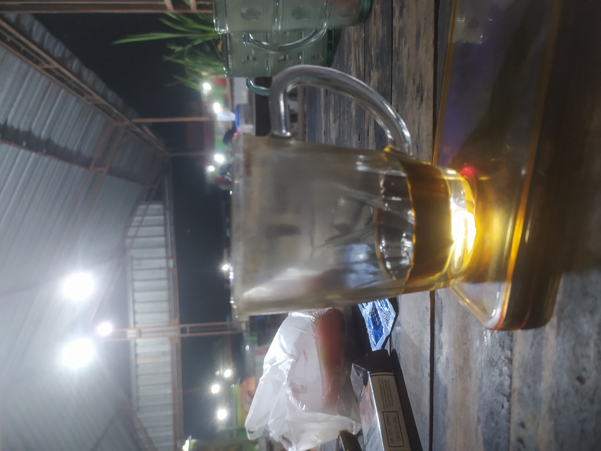 The image depicts a dimly lit outdoor seating area, likely a roadside cafe or warung, with a corrugated metal roof supported by wooden beams. Bright fluorescent lights illuminate the scene from above. On a rough wooden table in the foreground, a partially filled glass mug of dark liquid, possibly tea or coffee, sits on a transparent amber-colored placemat. To the left of the mug, a pack of cigarettes and a small blue packet are visible. Behind the mug, a white plastic bag contains a red, oval-shaped object, possibly fruit. In the background, other tables, chairs, and patrons are blurred, suggesting a casual gathering. The overall atmosphere is informal and nocturnal. The location is identified as Tuntang, Indonesia.