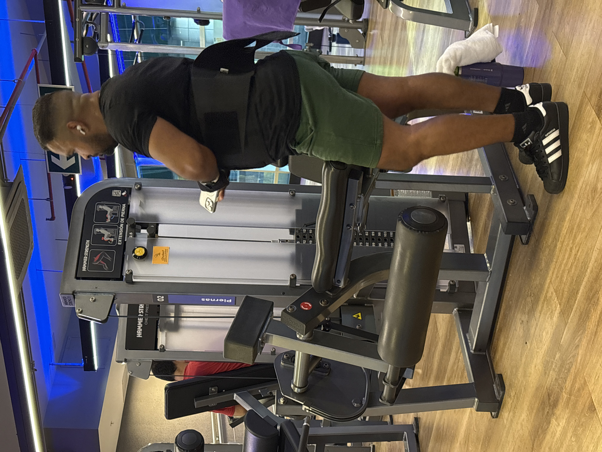 In a gymnasium located in San Isidro, Peru, a man with dark skin, a buzz cut, and a beard is standing near a weight machine. He is wearing a black t-shirt, green shorts, black socks, and black Adidas sneakers. A black weightlifting belt is cinched around his waist. In his right hand, he holds a smartphone. White wireless earbuds are visible in his ears. The weight machine is silver and grey, with black padded attachments. A diagram on the machine shows instructions for leg extensions. To the left of the man, another person wearing a red shirt is partially visible, also near a weight machine. The gym has a polished wooden floor and bright overhead lighting, with blue LED lights accentuating the ceiling. A white towel is draped over a blue water bottle positioned on the floor near the man's feet.