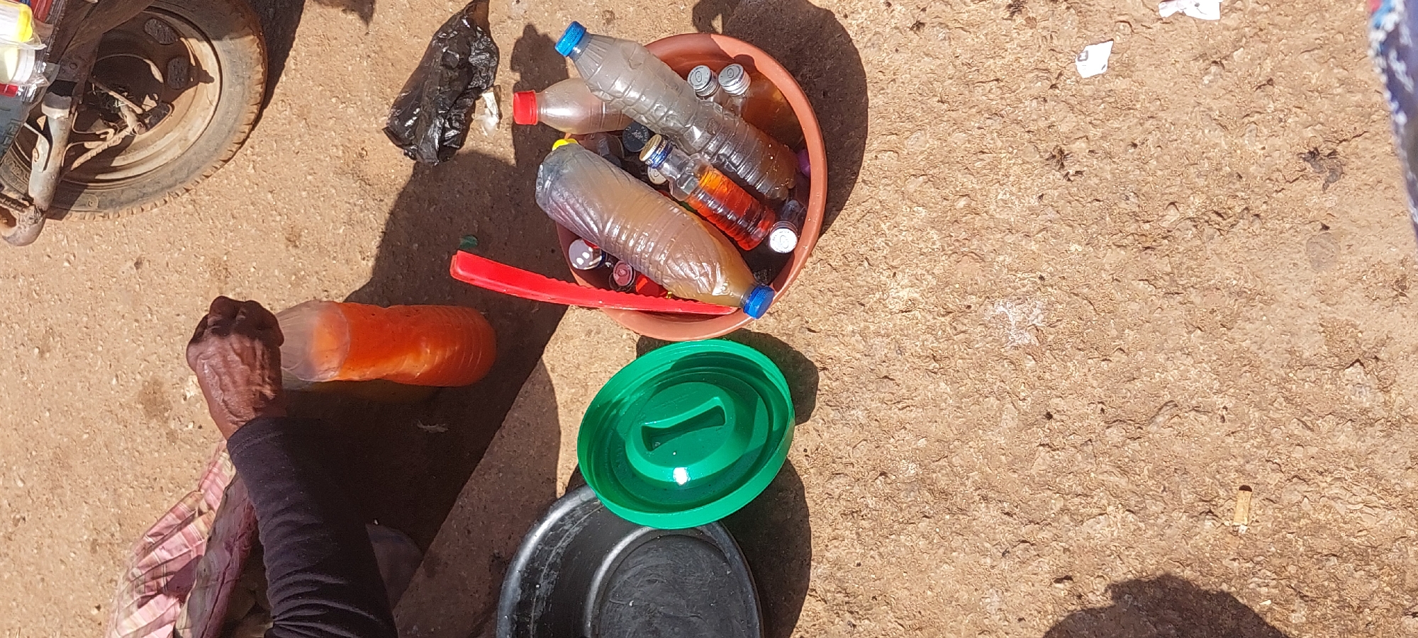 A close-up, overhead shot captures a collection of plastic bottles and containers on a dusty, uneven surface, likely soil or dirt. A person's wrinkled, dark-skinned hand is visible on the left, holding an orange-colored liquid in a clear plastic bottle. To the left of the hand, a black bowl and a bright green plastic lid are placed on the ground. A shallow brown bowl is filled with several clear plastic bottles containing various liquids, some appearing yellowish, amber, or clear, along with a red plastic object and a crumpled black plastic bag. In the background, the tire and part of the frame of a light-colored motorized vehicle are visible. The overall scene suggests an outdoor setting, possibly a market or roadside stall, where liquids are being sold or stored in reused plastic containers.