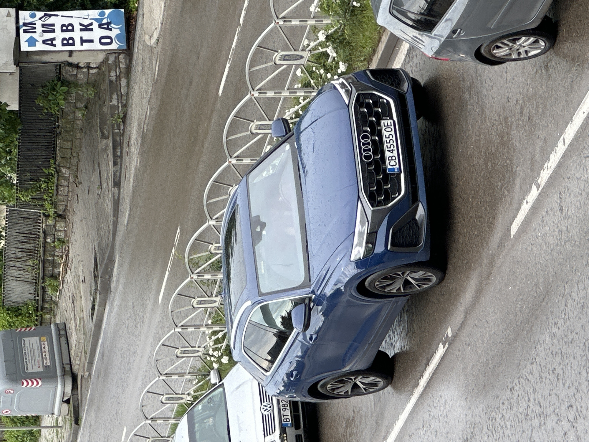 An elevated view captures a street scene, likely in Veliko Tarnovo, Bulgaria, featuring several parked cars on a damp asphalt surface, suggesting recent rain or overcast weather.
In the foreground, a prominent blue Audi SUV, possibly a Q3 or Q5 model, is parked, displaying a Bulgarian license plate "CB 4555 OE" (CB designates Sofia region, but the context states Veliko Tarnovo). Its sleek design and large grille with the Audi rings are clearly visible. Behind it, a silver or grey car is partially visible. In front of the Audi, a white car, identifiable by a Volkswagen logo on its grille, also has a Bulgarian license plate, reading "BT 982" (BT designates Veliko Tarnovo region).
Along the side of the road, a series of distinctive white, semi-circular metal barriers create a separation, behind which green plants and small white flowers grow. A concrete sidewalk or curb runs parallel to the road. On the far left, a large grey industrial-style trash bin or container with red and white striped markings is visible. Further left, a stone wall or building structure is present.
A blue and white sign, mounted on a building or wall, is partially visible on the upper left. The Cyrillic text on the sign reads "М АИ ВВ ТК ОА" with an arrow pointing upwards. This likely spells out "АВТОМИВКА" (AVTOMIVKA), meaning "car wash," indicating a nearby service in that direction. No people are visible, and the scene depicts a static moment of parked vehicles and urban infrastructure during the daytime.