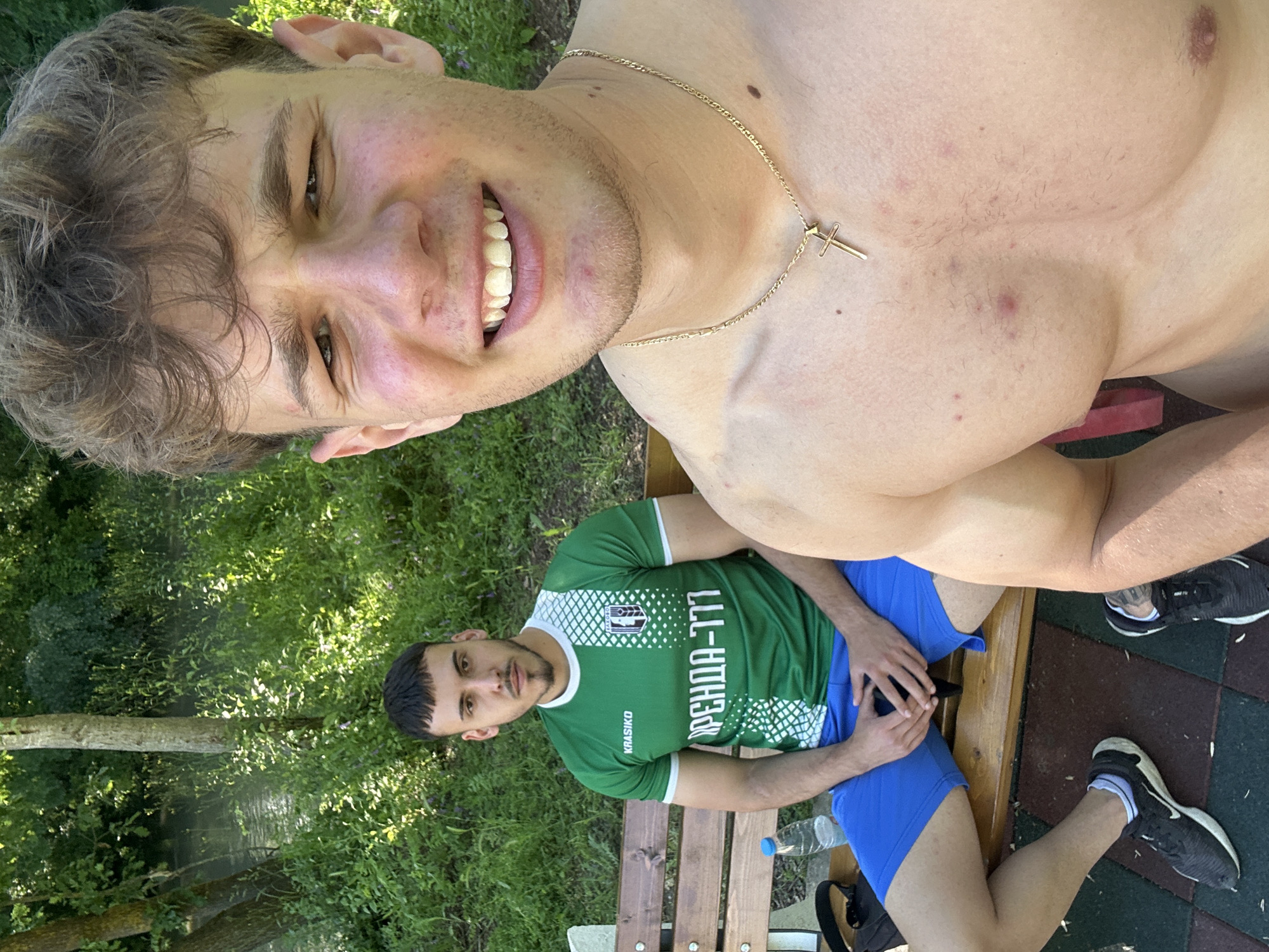 Two men are sitting on a wooden bench outdoors. One man is shirtless and wearing a gold chain with a cross pendant. He is smiling and appears to be taking a selfie. The other man is wearing a green and white soccer jersey, blue shorts, and sneakers. A water bottle sits on the bench beside him. The background includes trees, foliage, and what appears to be a body of water. The scene is set outdoors, likely a park or recreational area. The city is Elhovo, Bulgaria, based on the Cyrillic text on the jersey.