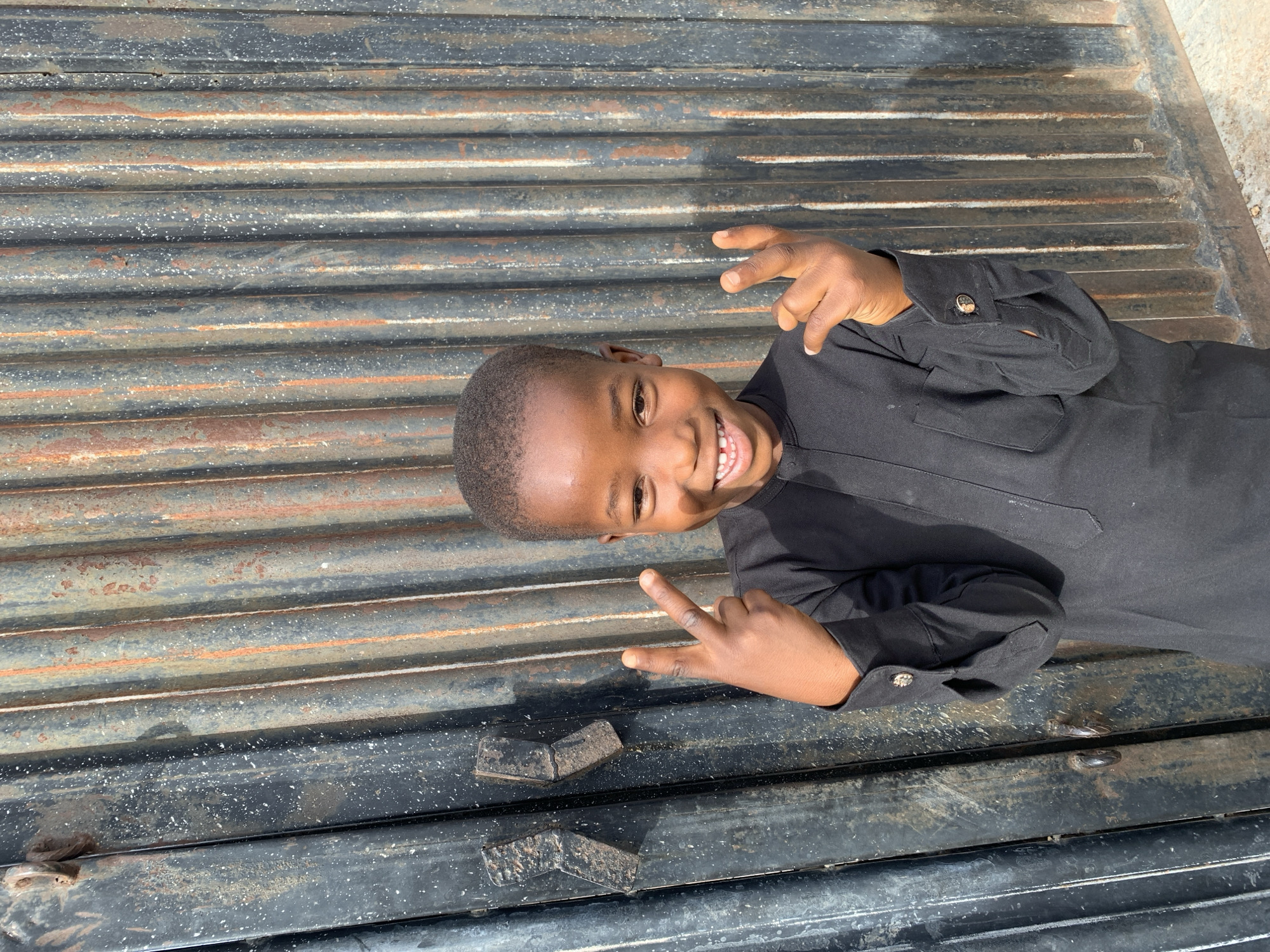 A young child is lying on a corrugated metal surface. The child is dark-skinned and wears a black long-sleeved garment, and is smiling. The child is also holding up two fingers in a "peace" sign. The metal surface is dark with streaks of rust and dirt. The scene takes place during daylight hours. While the location of the scene is unspecified in the image, the prompt asks the scene takes place in the city of Jos, Nigeria.