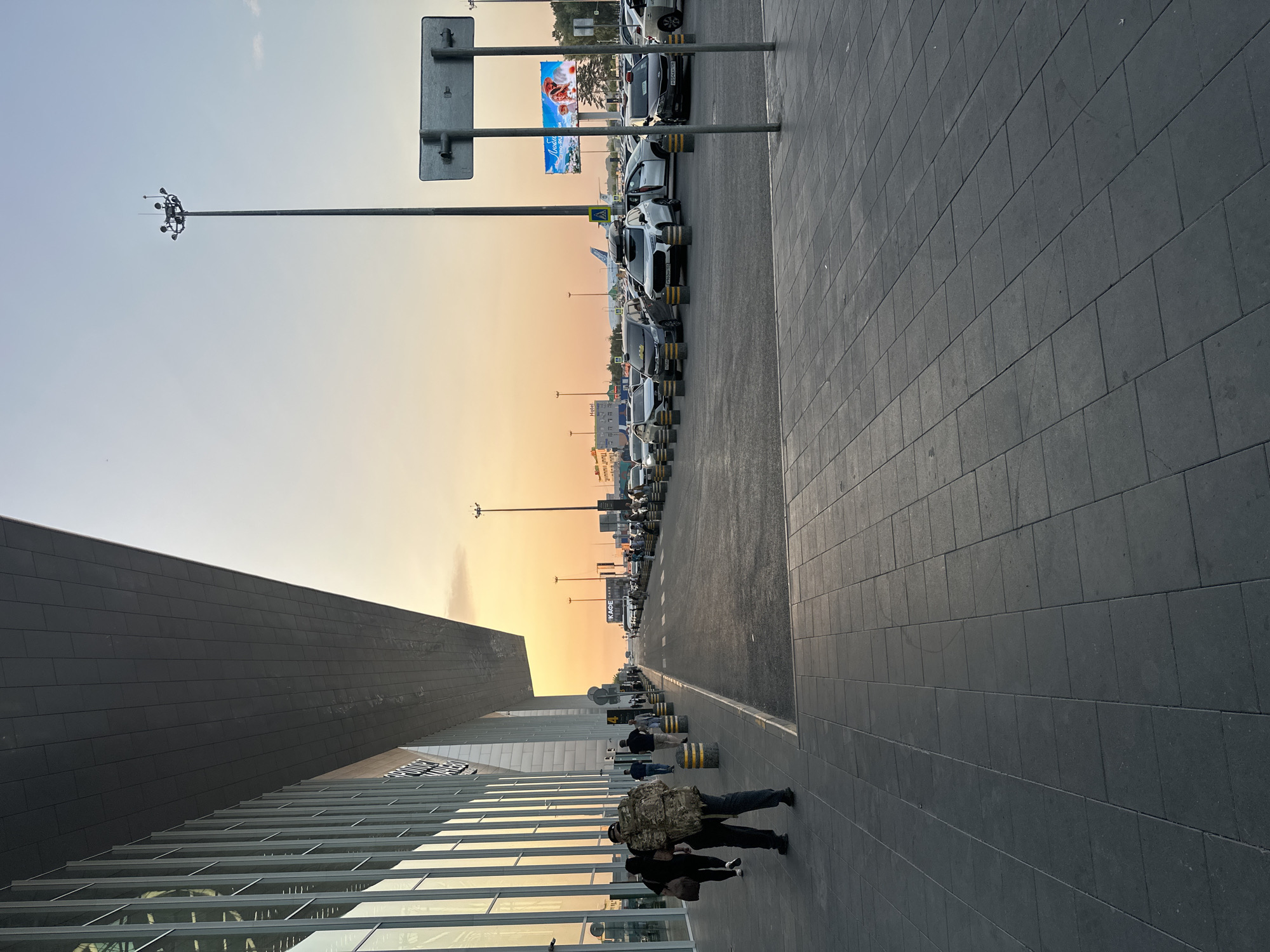 An exterior shot at sunset captures an airport terminal with a modern, angular design. The building features a large glass facade and a dark, cantilevered roof. A paved walkway runs alongside the terminal, with several people walking and standing. Beyond the walkway is a parking lot filled with vehicles, including cars and what appear to be taxis. In the background, several airplanes are visible on the tarmac, along with a tall light pole and a large billboard. The sky displays warm hues of orange and yellow, indicative of the setting sun. The ground is paved with dark gray square tiles.