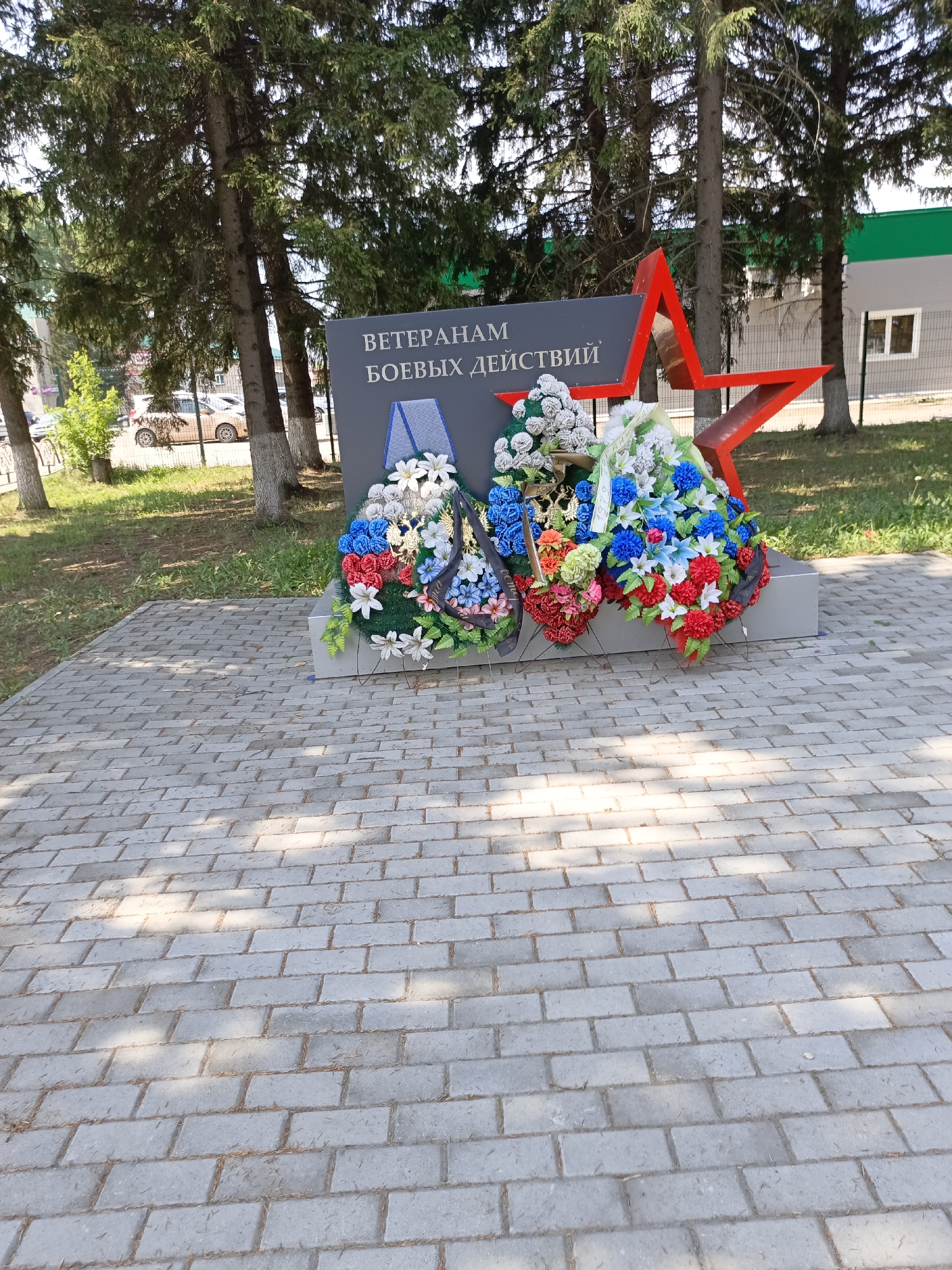 This image depicts a memorial monument in Zalesovo, Russia, dedicated to veterans of combat operations. The monument features a dark gray slab with the inscription "ВЕТЕРАНАМ БОЕВЫХ ДЕЙСТВИЙ" (To Veterans of Combat Operations) in white capital letters. A red five-pointed star is affixed to the right side of the slab. In front of the monument are two artificial flower wreaths, one predominantly white and blue, and the other a mix of red, blue, and white flowers, with a ribbon bearing Cyrillic text that is partially obscured. The monument is situated on a paved walkway made of grey rectangular bricks. Behind the monument, several tall pine trees are visible, casting shadows on the ground. In the background, there is a building with a green and gray facade and a white-framed window, as well as a fence and some parked vehicles. The scene is outdoors, suggesting it is daytime, with sunlight filtering through the trees.