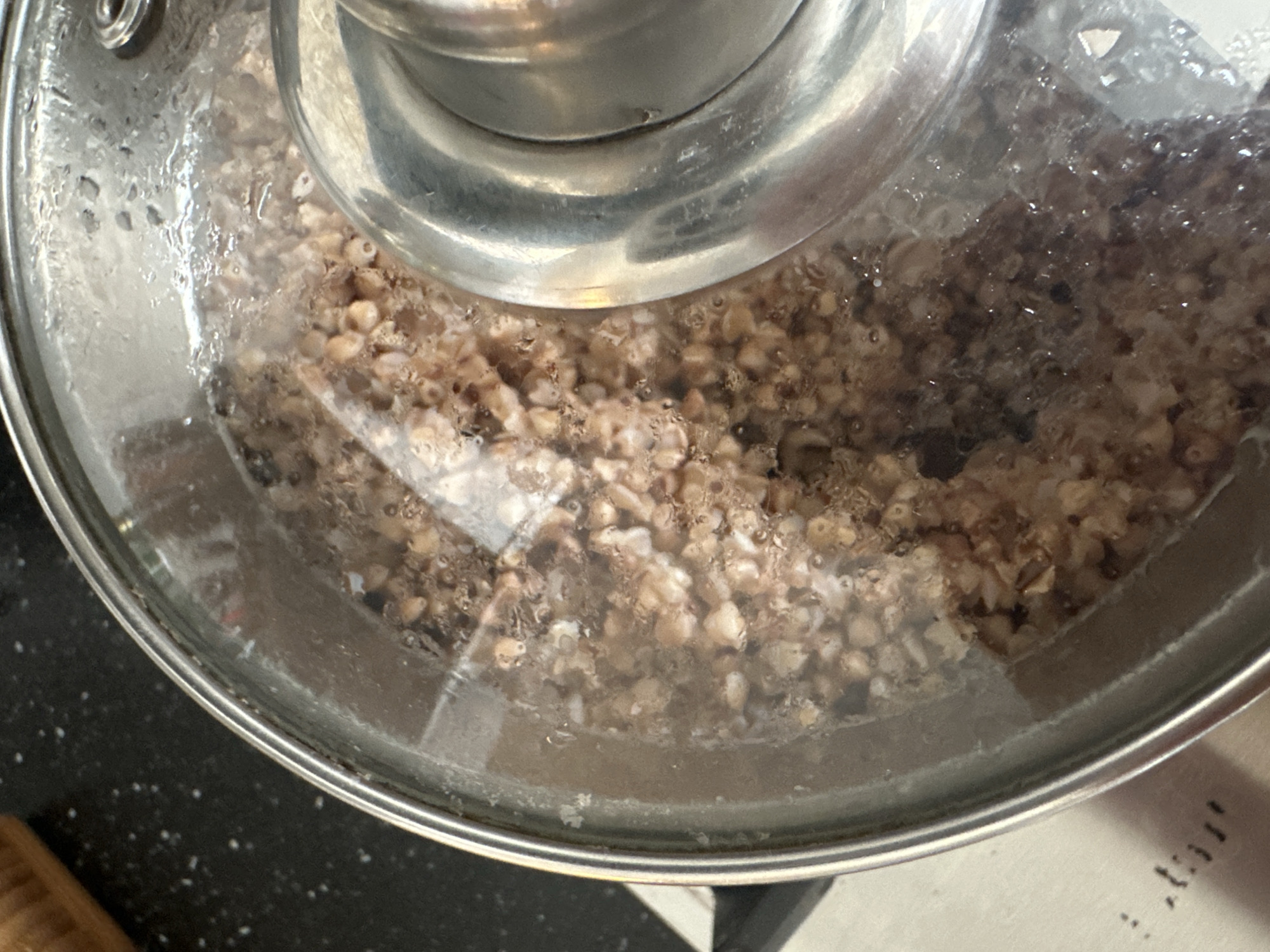 The image shows a close-up, overhead view of buckwheat being cooked in a pot with a clear glass lid. The buckwheat grains are visible through the condensation on the lid, and the pot appears to be on a stovetop. The background shows a dark, speckled countertop, possibly granite. There is also a portion of a wooden rolling pin or similar kitchen utensil visible in the upper left corner. The scene is indoors and appears to be in a kitchen setting. The location in the city of Nevinnomyssk, Russia, cannot be determined from the provided image.