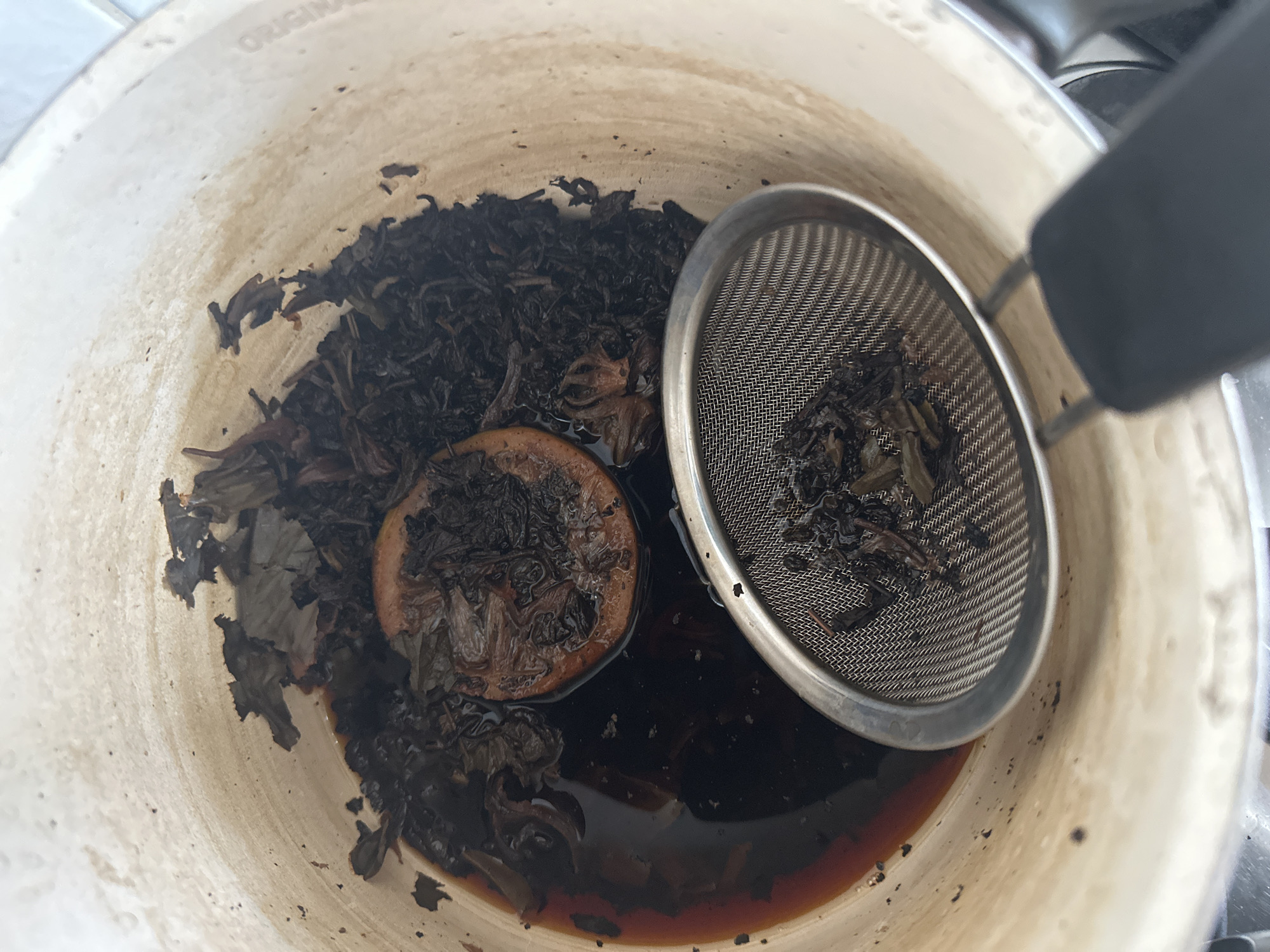 A close-up, top-down view shows a pot containing dark brown liquid, loose tea leaves, and a slice of dried citrus, possibly orange, in Nevinnomyssk, Russia. A metal mesh tea strainer with some tea leaves is partially submerged in the liquid on the right side of the pot. The interior of the pot appears to have some residue or staining. The focus is on the contents of the pot, with the rim and the exterior of the pot slightly visible at the top and right edges.