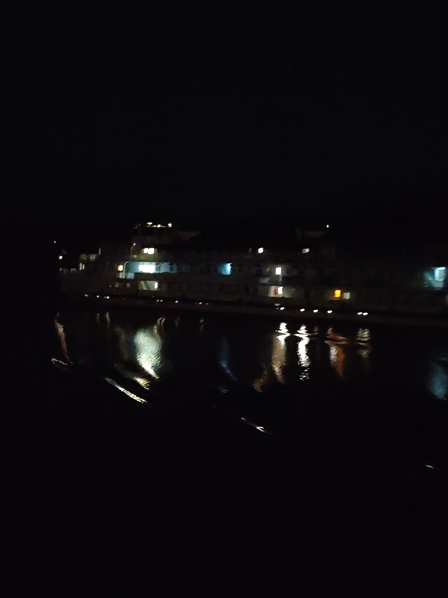 The image depicts a body of water at night, possibly a river or lake, reflecting the lights of a multi-story building on the opposite bank. The building has multiple illuminated windows and appears to be a large structure. The scene suggests an urban or semi-urban environment at night, and the presence of the water implies a location near a riverbank.