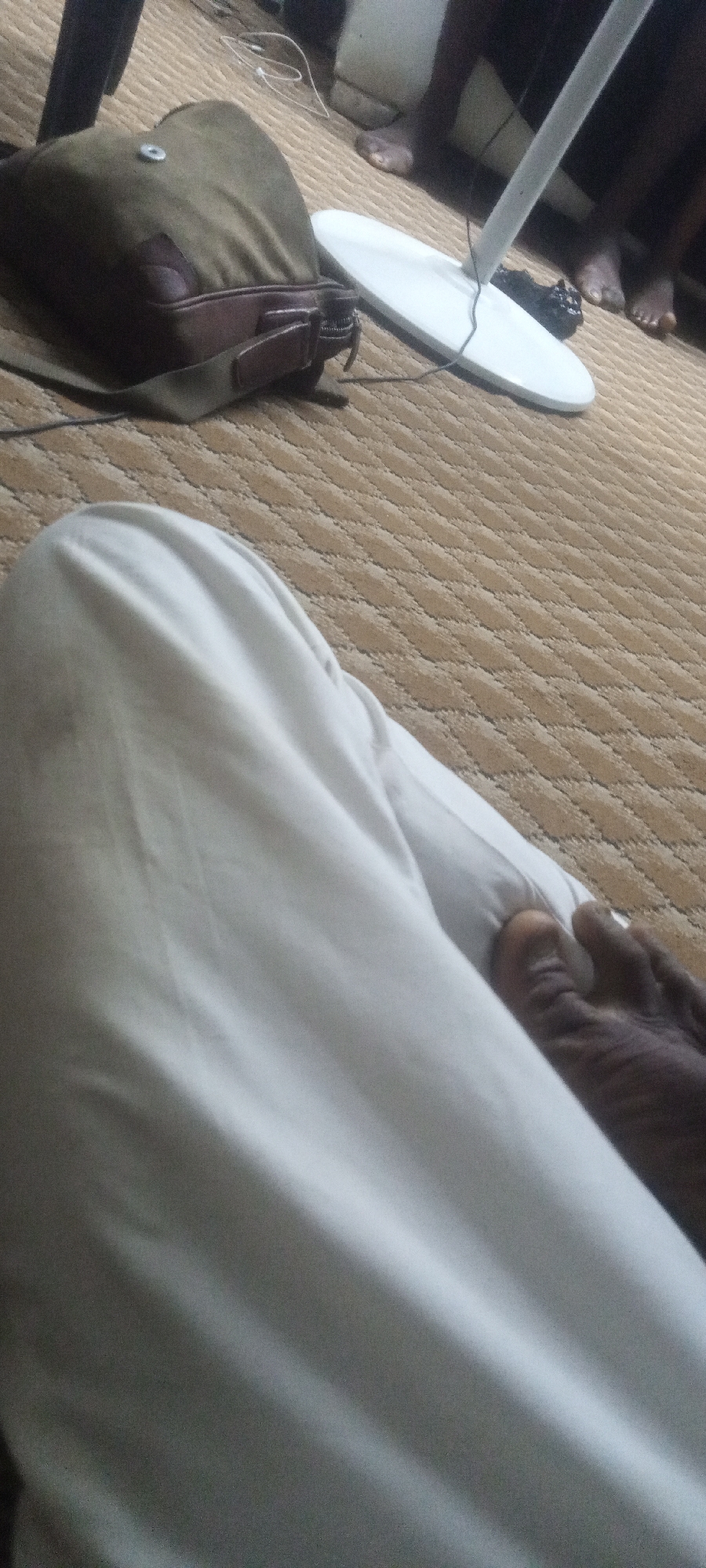The image captures a close-up perspective of a person reclining, with their lower body visible, set against a backdrop of a carpeted interior.
A leg, covered in white fabric, occupies the foreground, with the toes of a dark-skinned foot peeking out from under the fabric. To the left and further back, there's a brown bag with a strap resting on the carpet. In the background, partially obscured, is a white circular object, possibly the base of a lamp or stand, and glimpses of other people's feet. The carpet has a patterned texture. The location in the city of Unknown, Unknown. is unknown.