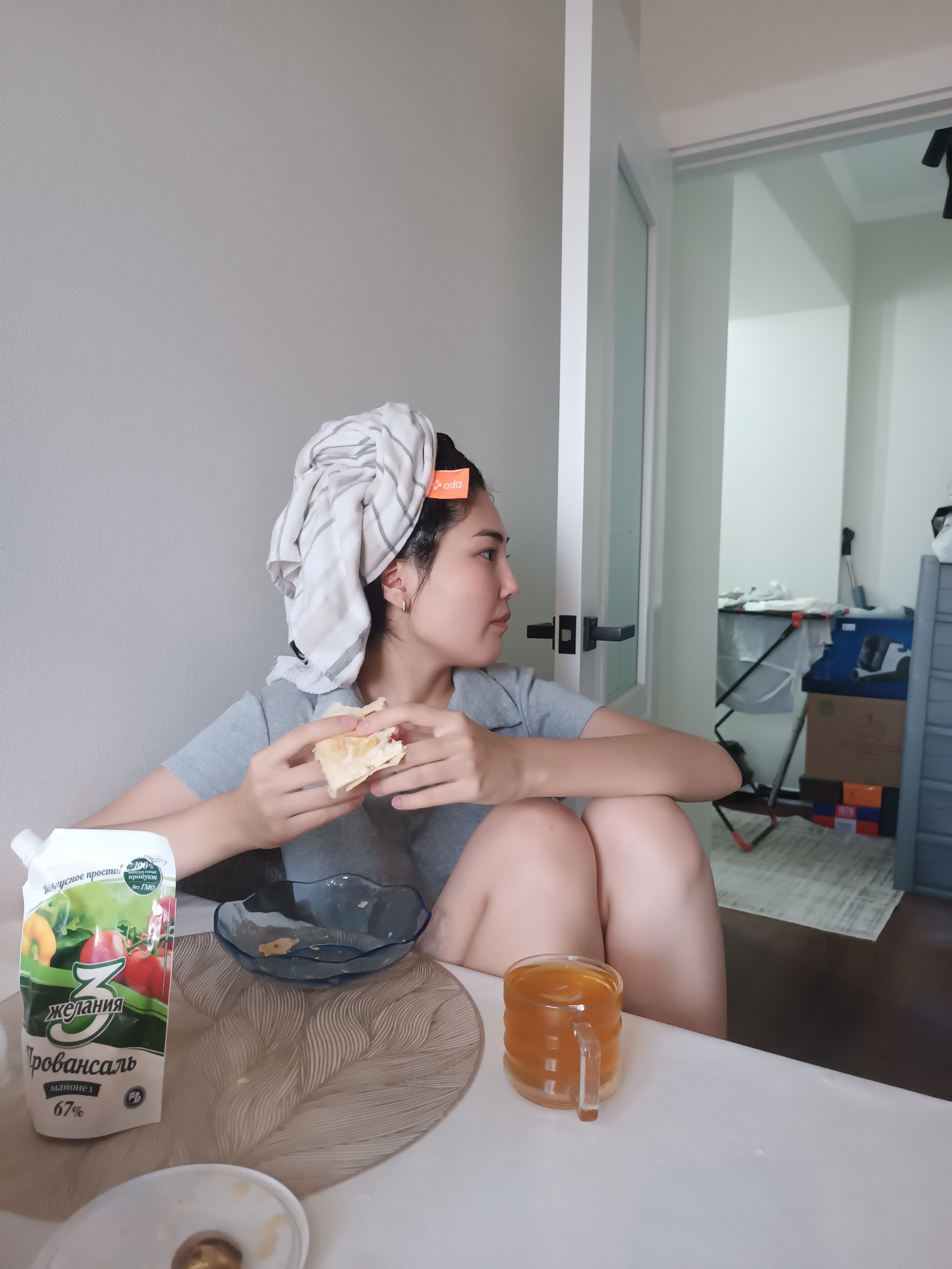 A woman with a towel wrapped around her head is seated at a table, holding a piece of food in her hands and looking to the side. She is wearing a gray t-shirt. On the table in front of her are a pouch of mayonnaise, a blue bowl, and a glass mug filled with a golden liquid. The background shows a doorway leading to another room with boxes and a laundry basket. The lighting is natural and comes from the side.