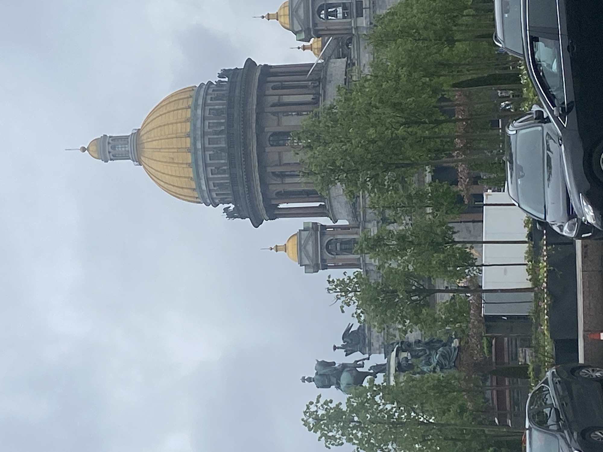 The image captures St. Isaac's Cathedral in Saint Petersburg, Russia. The cathedral's golden dome is prominently visible against a cloudy sky. The equestrian statue of Nicholas I stands in the foreground. Trees line the street. Modern cars are parked along the road, positioned in the right of the frame.