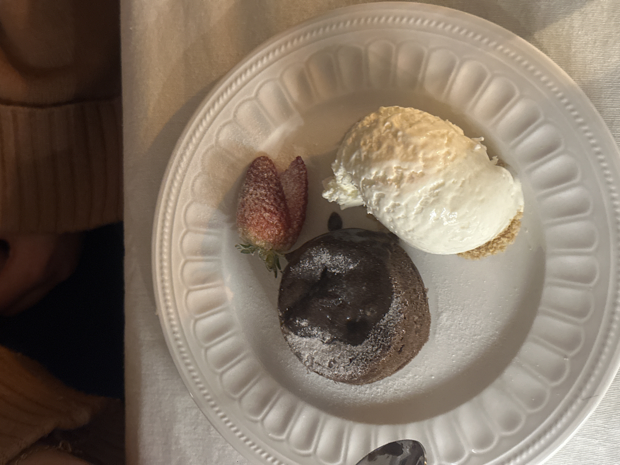 A white plate is viewed from above, holding a dessert consisting of a chocolate lava cake dusted with powdered sugar, a scoop of white ice cream, and two strawberry halves. A spoon is partially visible at the bottom of the plate. The plate sits on a white tablecloth. The edge of a chair or a person's clothing is visible on the left side of the image. There is no information available to suggest that this image was taken in Yerevan, Armenia.