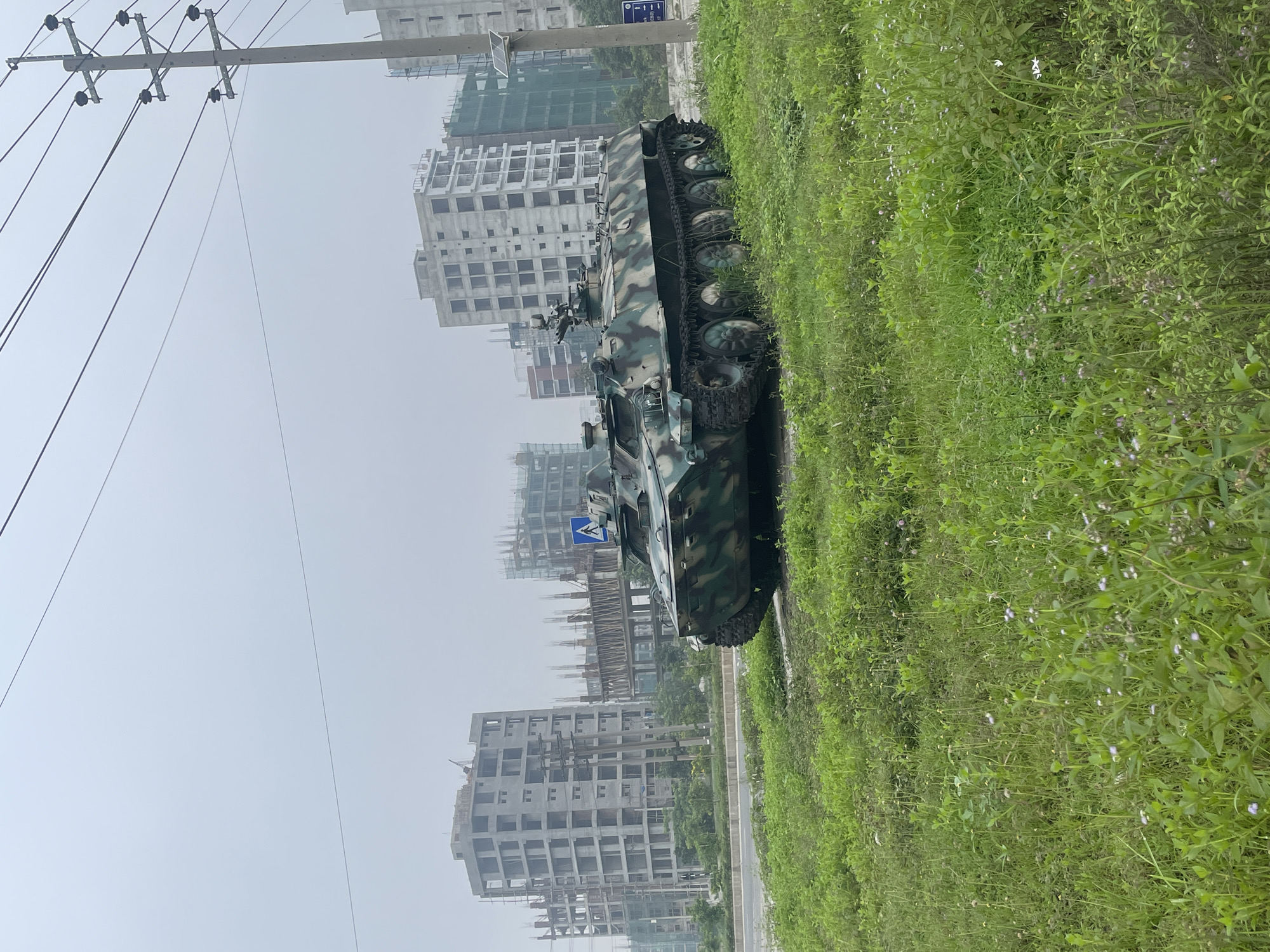 The image shows a military vehicle, possibly an armored personnel carrier, parked on the side of a road in an urban area. The vehicle is camouflaged and positioned on the grass next to the road. In the background, there are several multi-story buildings, some of which appear to be under construction. Utility poles with wires are visible on the left side of the image. The sky is overcast. Based on the prompt, the location is Rupganj, Bangladesh.