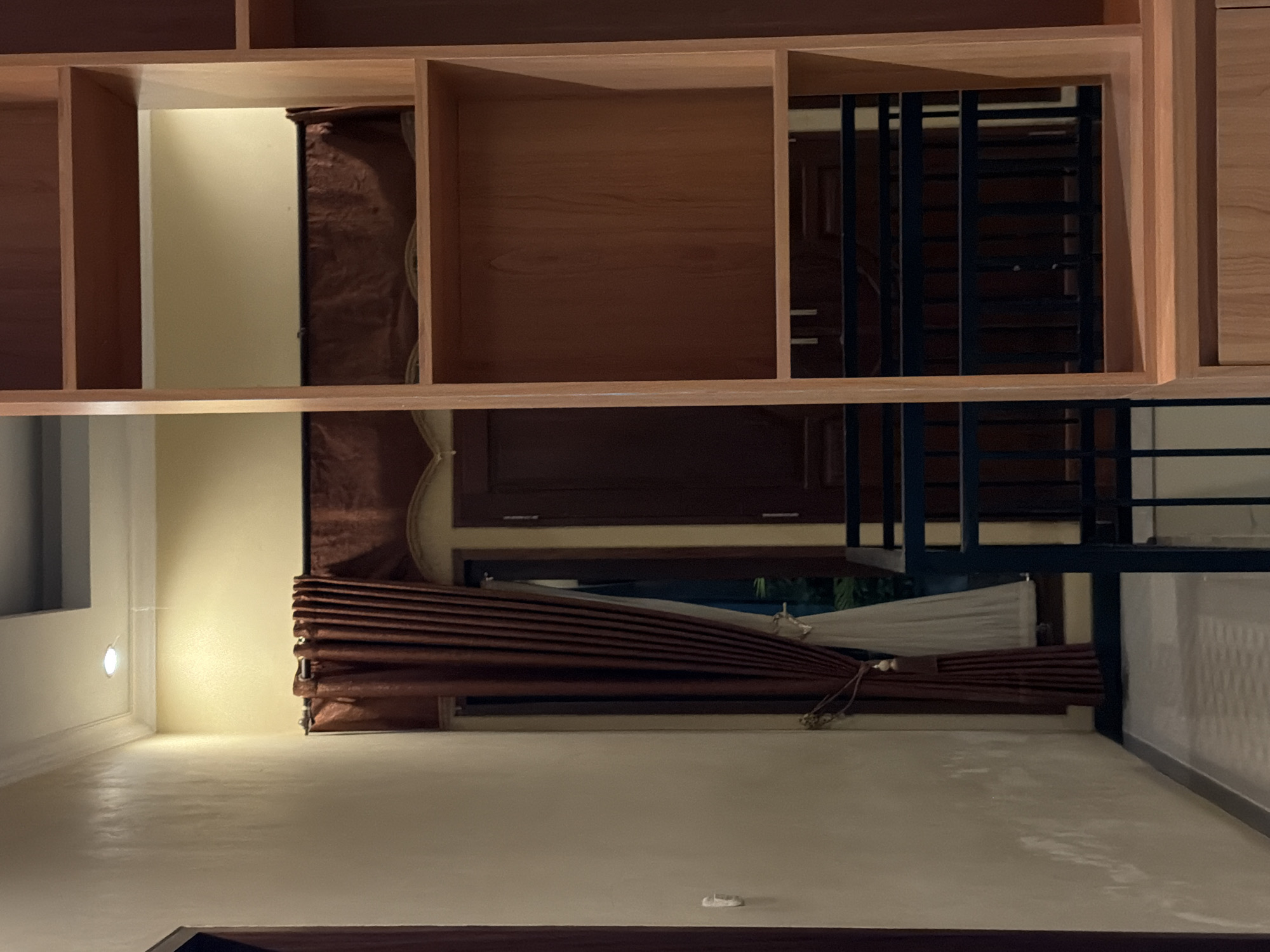 The image shows a corner of a room with a wall on the left and a tall, wooden bookshelf on the right. The wall is painted a light beige and has some texture. To the right of the wall, there is a doorway with a dark brown door and dark brown curtains. The curtains are drawn back and tied with a decorative tassel. Beyond the door, there appears to be a balcony or hallway with a black metal railing.

The bookshelf is made of light-colored wood and has several open shelves. The shelves are empty except for one in the middle which contains some dark fabric, possibly a blanket or cushion. The top of the bookshelf has a visible ceiling with a square recessed lighting fixture. The floor in the foreground is a light grey ramp with a textured surface. The lighting in the room is dim, with a single light source visible in the ceiling.

The scene appears to be an interior shot, possibly in a residential or commercial building. The style of the door and curtains suggests an Indonesian aesthetic. Without further context, it is impossible to determine the exact location within Wonoayu, Indonesia.