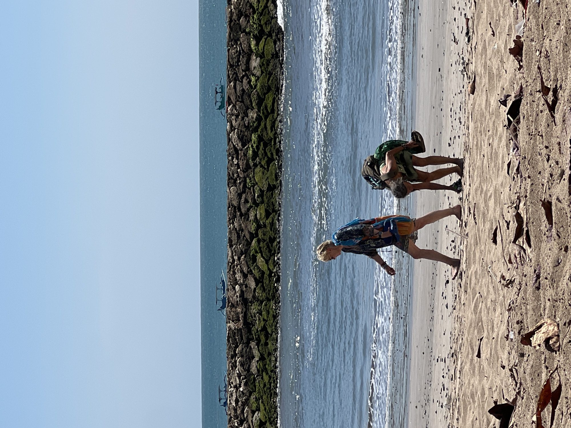 This image captures a scene on a beach in Kuta, Indonesia, with the ocean stretching out to the horizon. In the foreground, two individuals are present on the sandy shore. To the left, a person with blonde hair, wearing sunglasses, a patterned dress, and carrying a bag, walks along the beach. To the right, a second individual, with long dark hair and wearing a green garment, is bent over, appearing to pick something up from the sand. They are carrying a backpack and holding a pair of flip-flops. In the background, a stone breakwater runs parallel to the coastline, with moss-covered rocks. Beyond the breakwater, several traditional Balinese fishing boats with canopies are visible on the turquoise water. The sky is clear and blue. The beach itself is littered with natural debris, including dried leaves and twigs, as well as some man-made detritus.