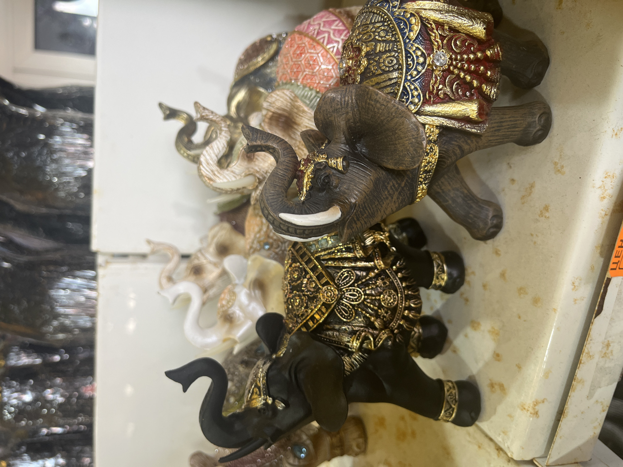 In the foreground, two ornate elephant figurines are displayed on a shelf. The figurine on the left is black with intricate gold detailing on its blanket and ankles. The figurine on the right is brown with a similar elaborate gold and red blanket and a small bell hanging from its trunk. Behind these two, several other elephant figurines in varying colors and designs are visible, some appearing to be made of a lighter material like ceramic or resin. The background consists of blurry shelves and objects, suggesting an indoor retail environment. The image does not contain any specific information or context related to the city of Plastunovskaya, Russia.