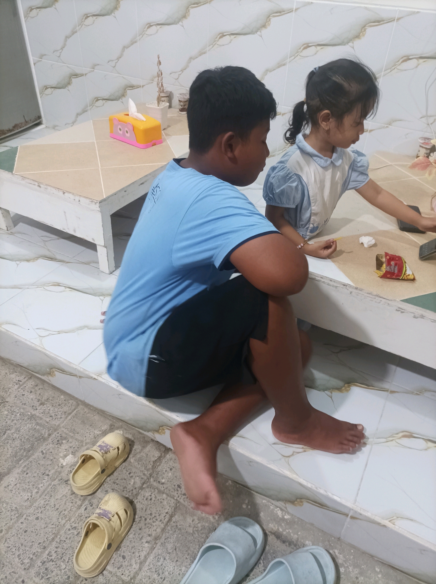 In Canga'an, Indonesia, two children are seated on a tiled surface. A boy in a blue t-shirt and black shorts is on the left, with his legs crossed. To his right, a girl in a light blue and white dress is also seated. They appear to be interacting with items on a table in front of them. On the table, there is a yellow tissue box with a cat design, a small potted plant, a bag of chips, and what appears to be a mobile phone. There are also two pairs of slip-on shoes visible on the floor in the foreground. The environment is indoors, with tiled walls and floor.