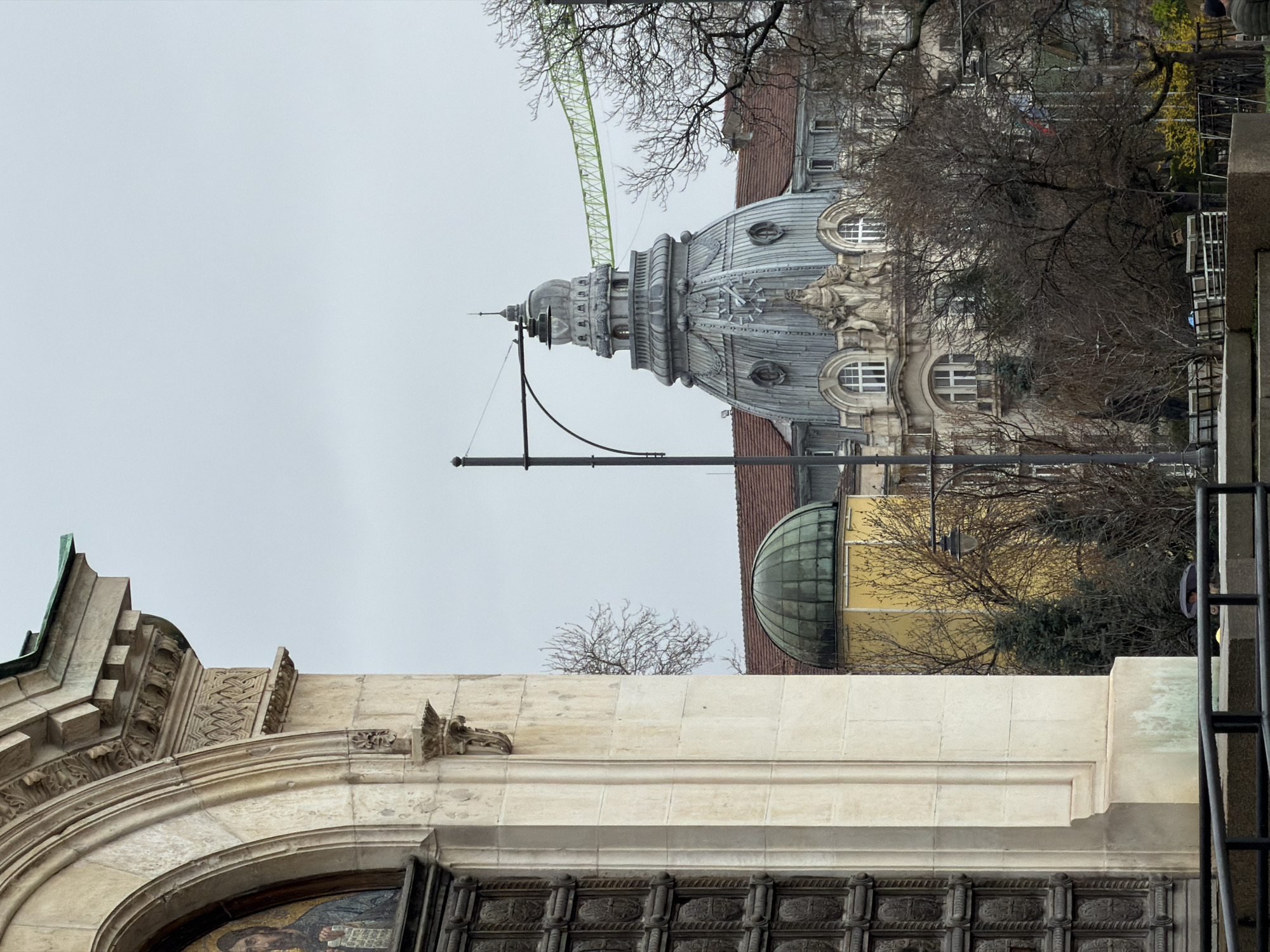 The image shows architectural details of buildings in Sofia, Bulgaria. A large, ornately decorated dome with a green crane in the background is visible. The lower portion of the image features a building with arched windows and detailed carvings. A domed building with a green roof is also partly visible. The sky is overcast.